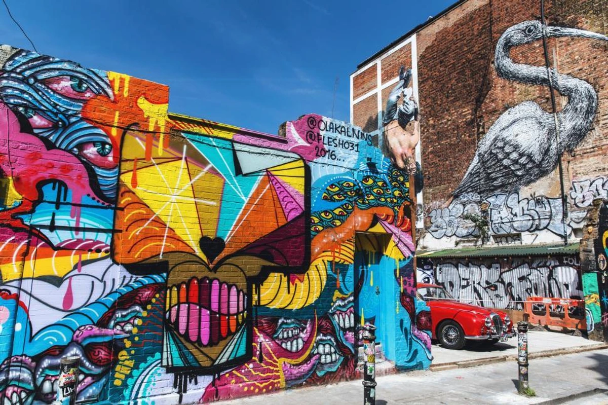Vibrant and colorful graffiti art covering brick walls in Brick Lane, London, featuring various styles and characters, including a large cat-like face and a heron, with a red vintage car parked nearby.