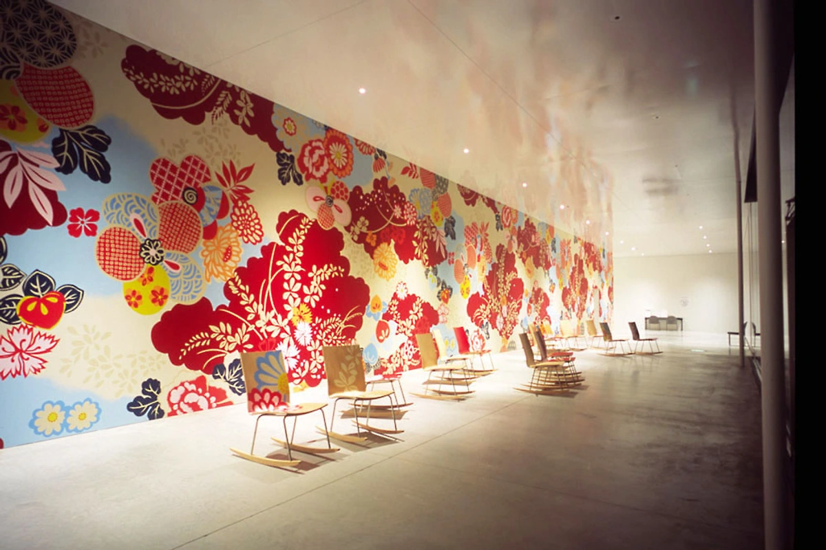 Installation at the 21st Century Museum of Contemporary Art in Kanazawa featuring rows of rocking chairs facing a large wall covered in colorful floral patterns.