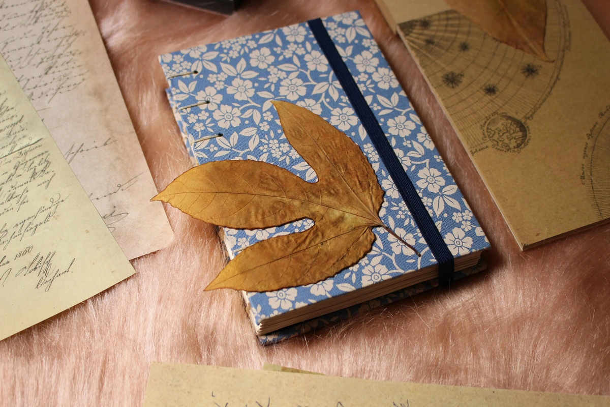 Aesthetic flat lay of junk journaling supplies including old letters, a floral notebook, and dried leaves on a pink textured surface.