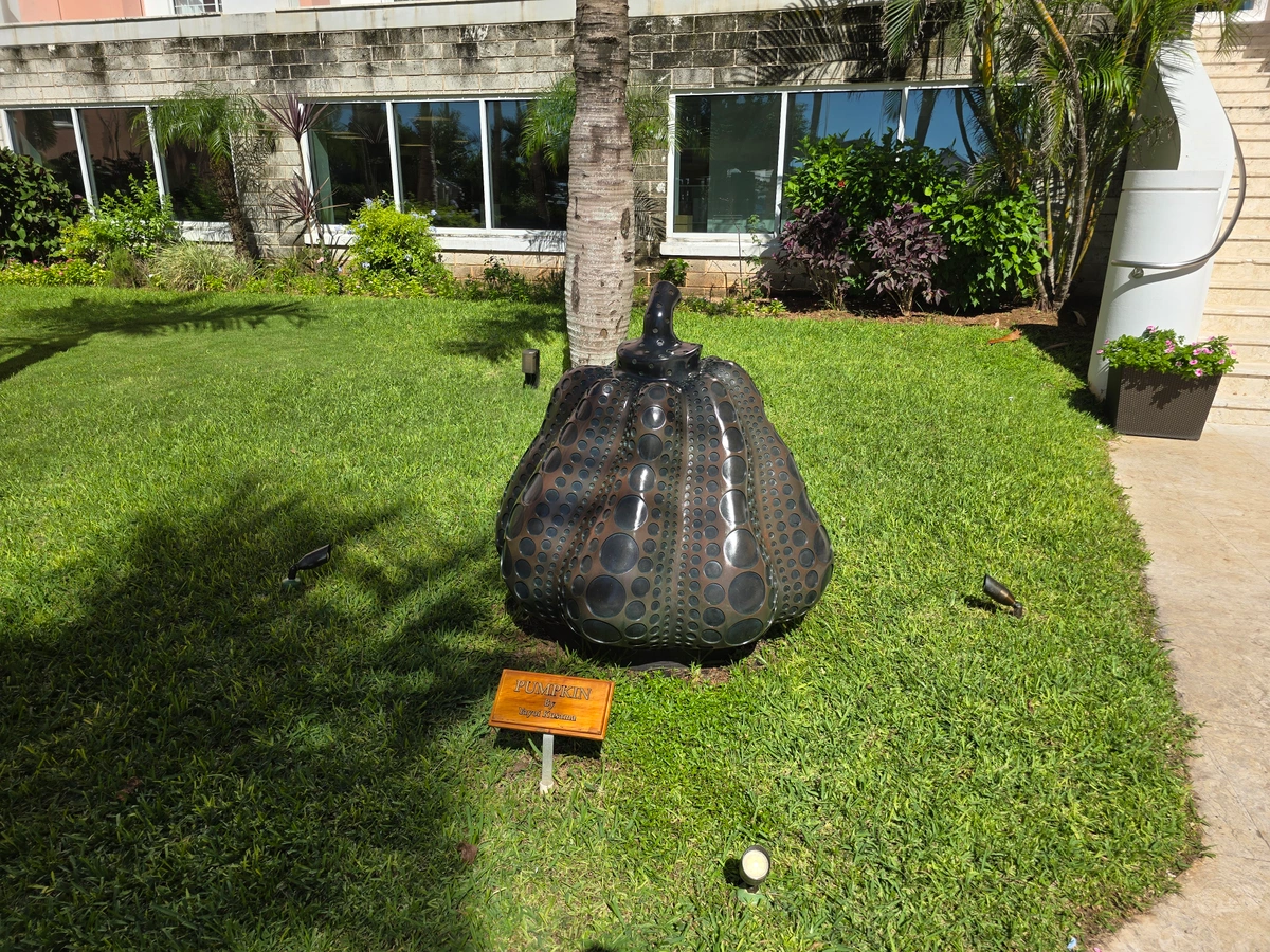 Yayoi Kusama's iconic 'Pumpkin' sculpture displayed outdoors on a green lawn, with a building and palm trees in the background.