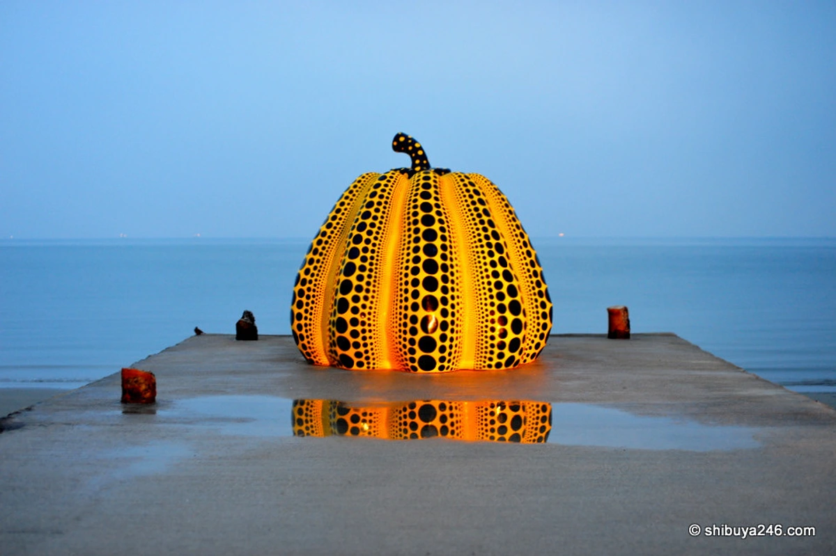 Naoshima's iconic yellow pumpkin sculpture by Yayoi Kusama on a pier at dusk, with its reflection in the water.