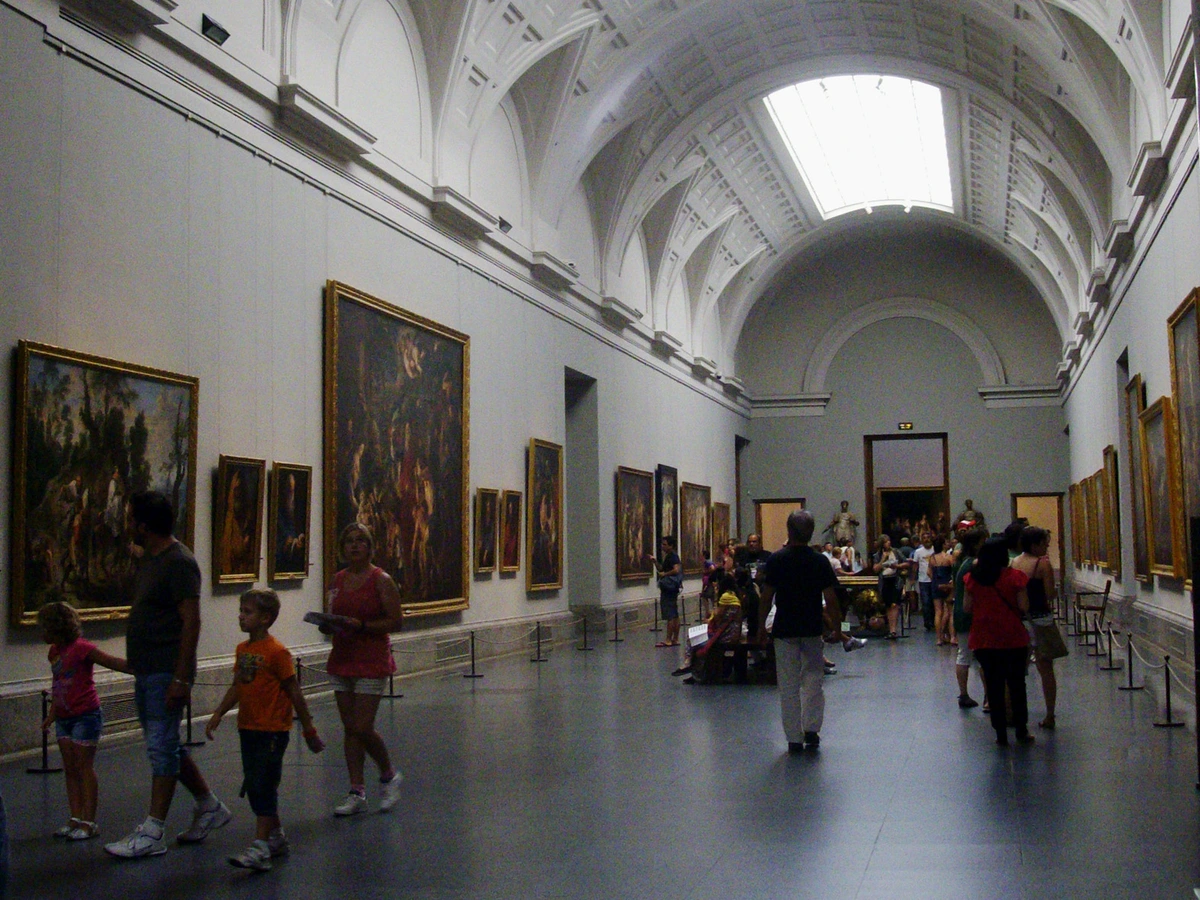 Interior view of the Prado Museum's permanent collection gallery with visitors viewing large, framed paintings under a high, arched ceiling with a skylight.