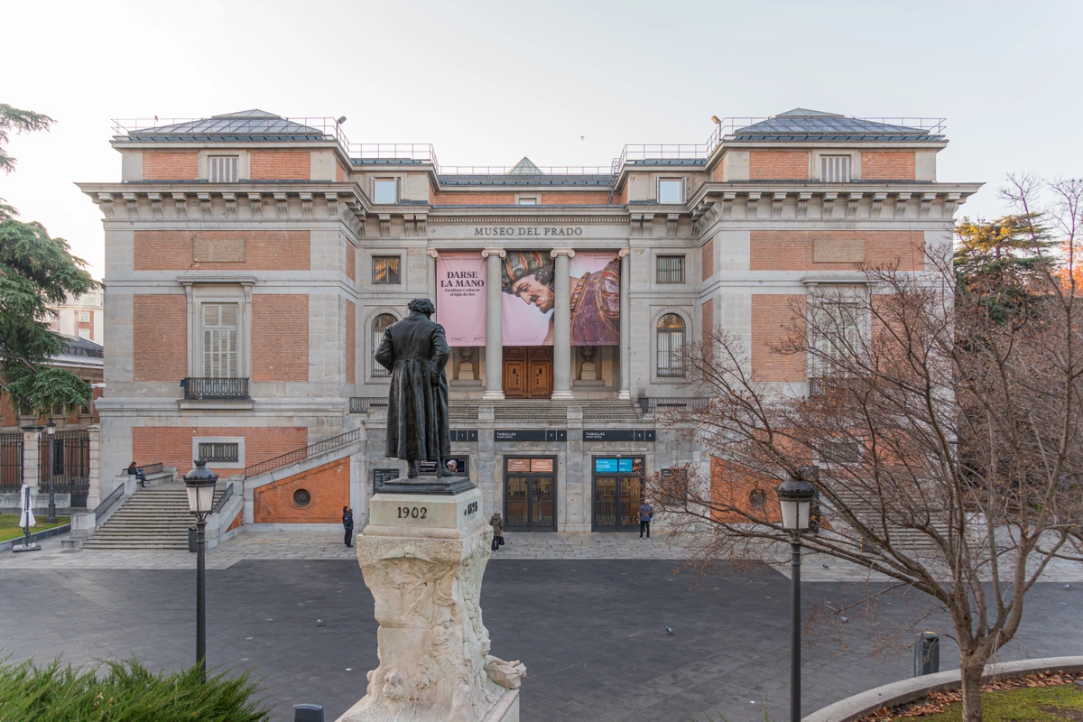 The main entrance of the Museo del Prado in Madrid, featuring a statue in the foreground and banners advertising an exhibition.