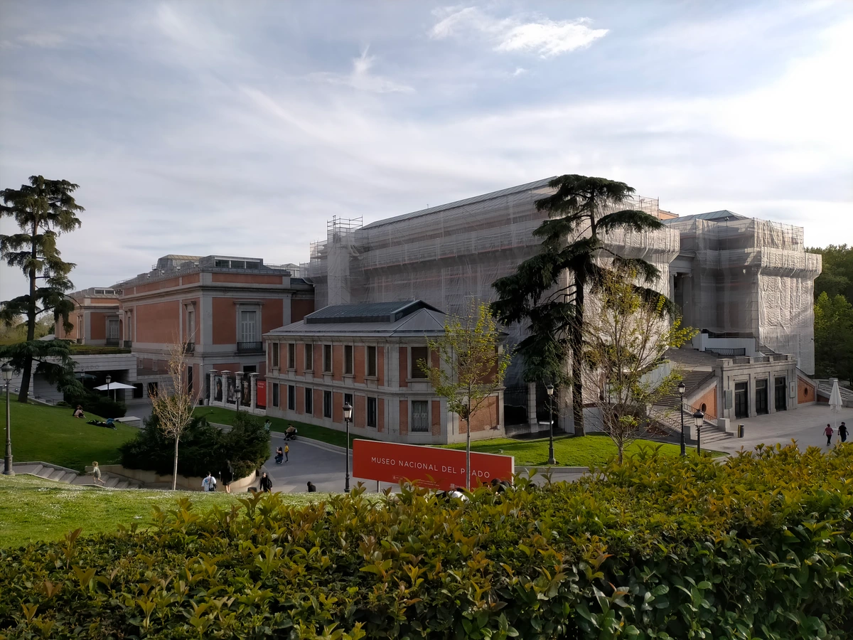 Exterior view of the Museo Nacional del Prado in Madrid, partially covered in scaffolding for renovations, with trees and a sign in front.