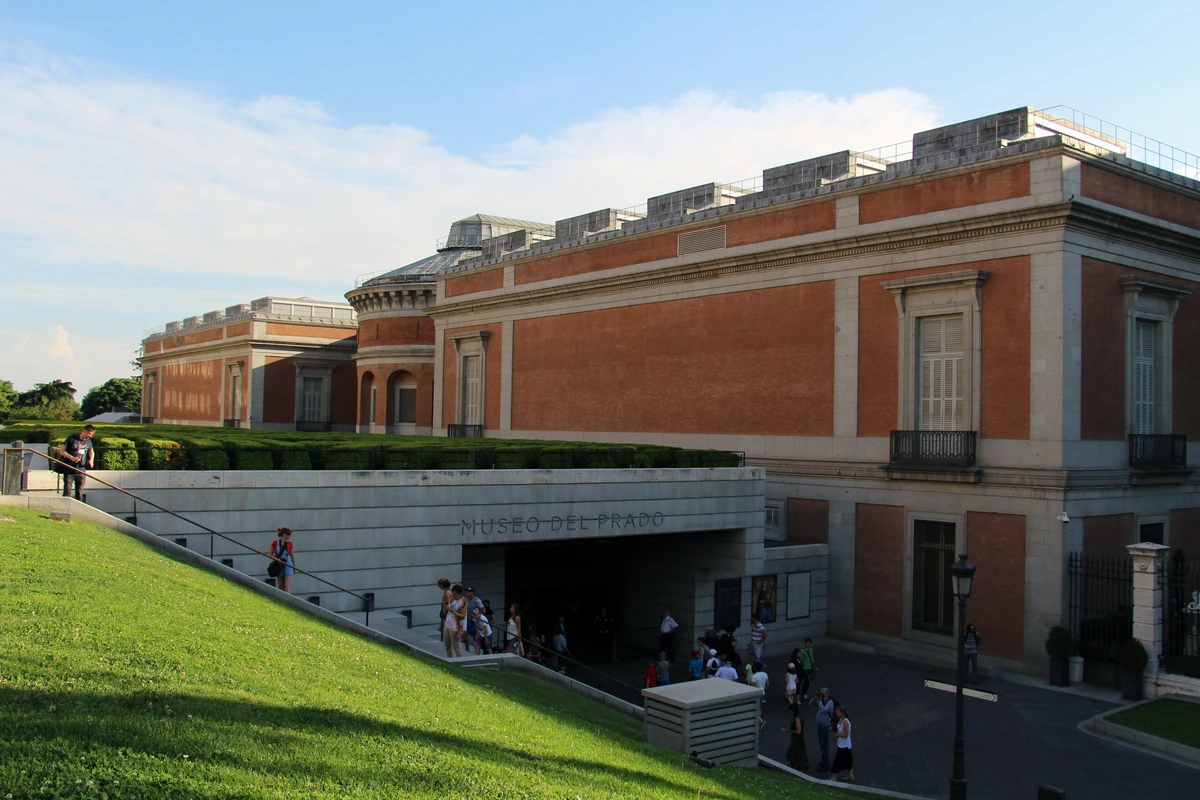 Exterior view of the Prado Museum entrance in Madrid, with people walking up the stairs and a grassy lawn in the foreground.
