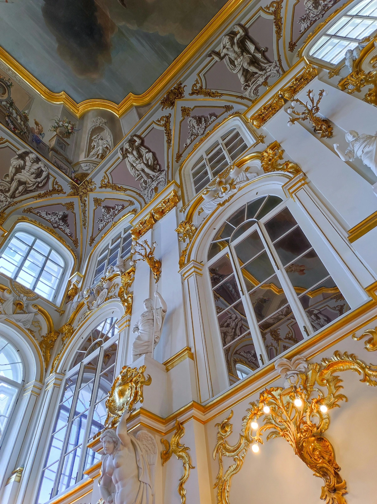Opulent interior of the Winter Palace in Saint Petersburg, Russia, featuring gilded ornamentation, frescoes, and large arched windows.