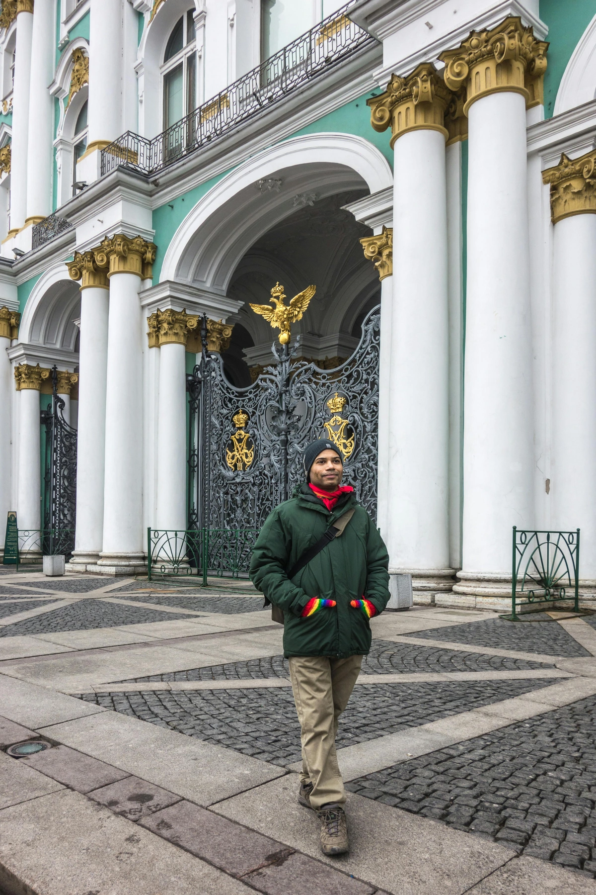 Man in a green jacket and rainbow gloves standing in front of the ornate gates of the Hermitage Museum in St. Petersburg, Russia.