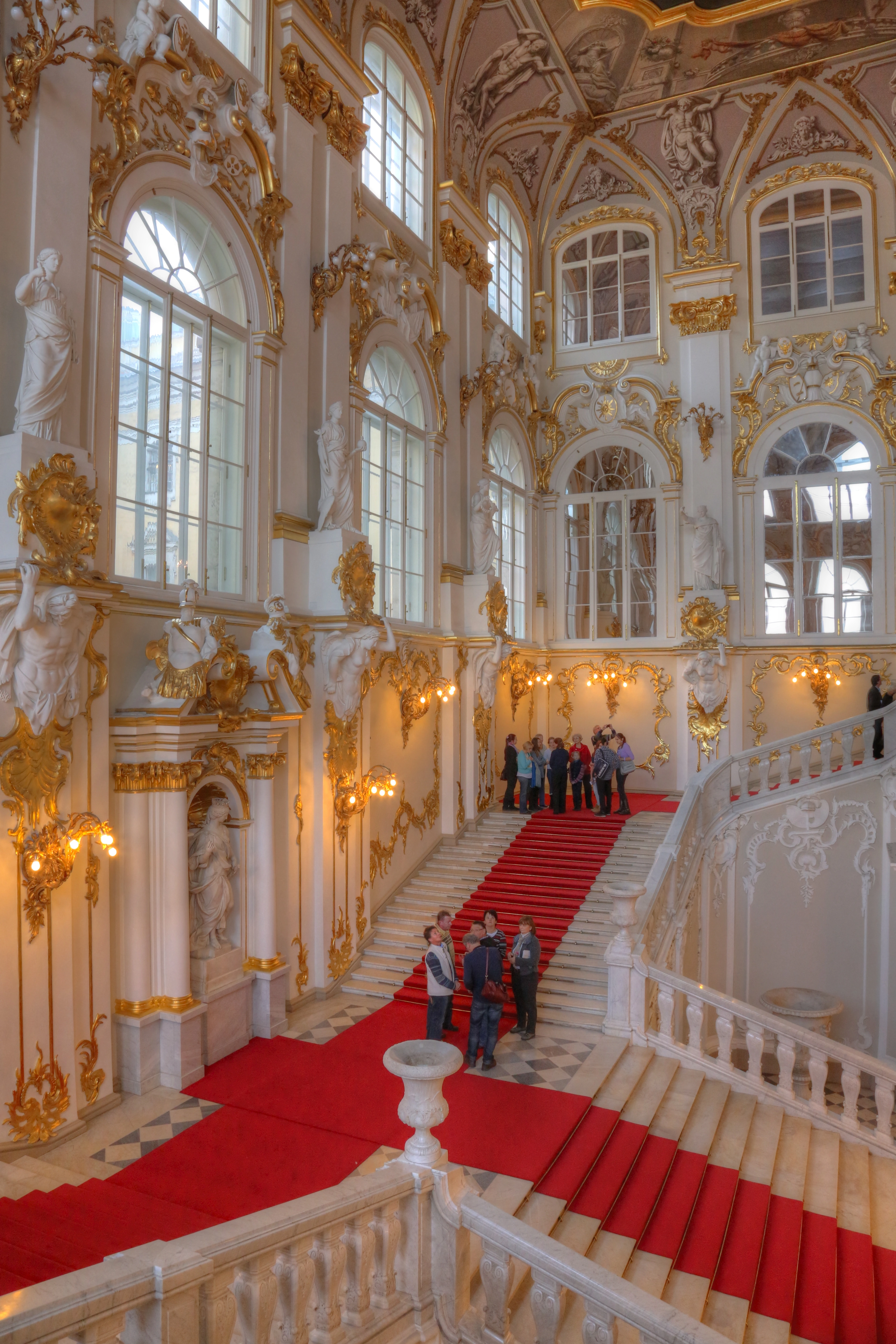 Grand staircase with red carpet inside the Hermitage Museum, St. Petersburg, Russia, adorned with ornate gilded decorations and sculptures.