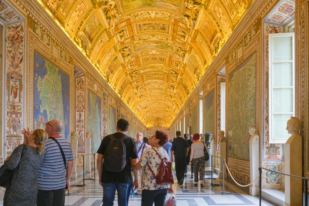 Visitors walk through a grand, ornate corridor in the Vatican Museums, admiring large map tapestries and richly decorated ceilings.