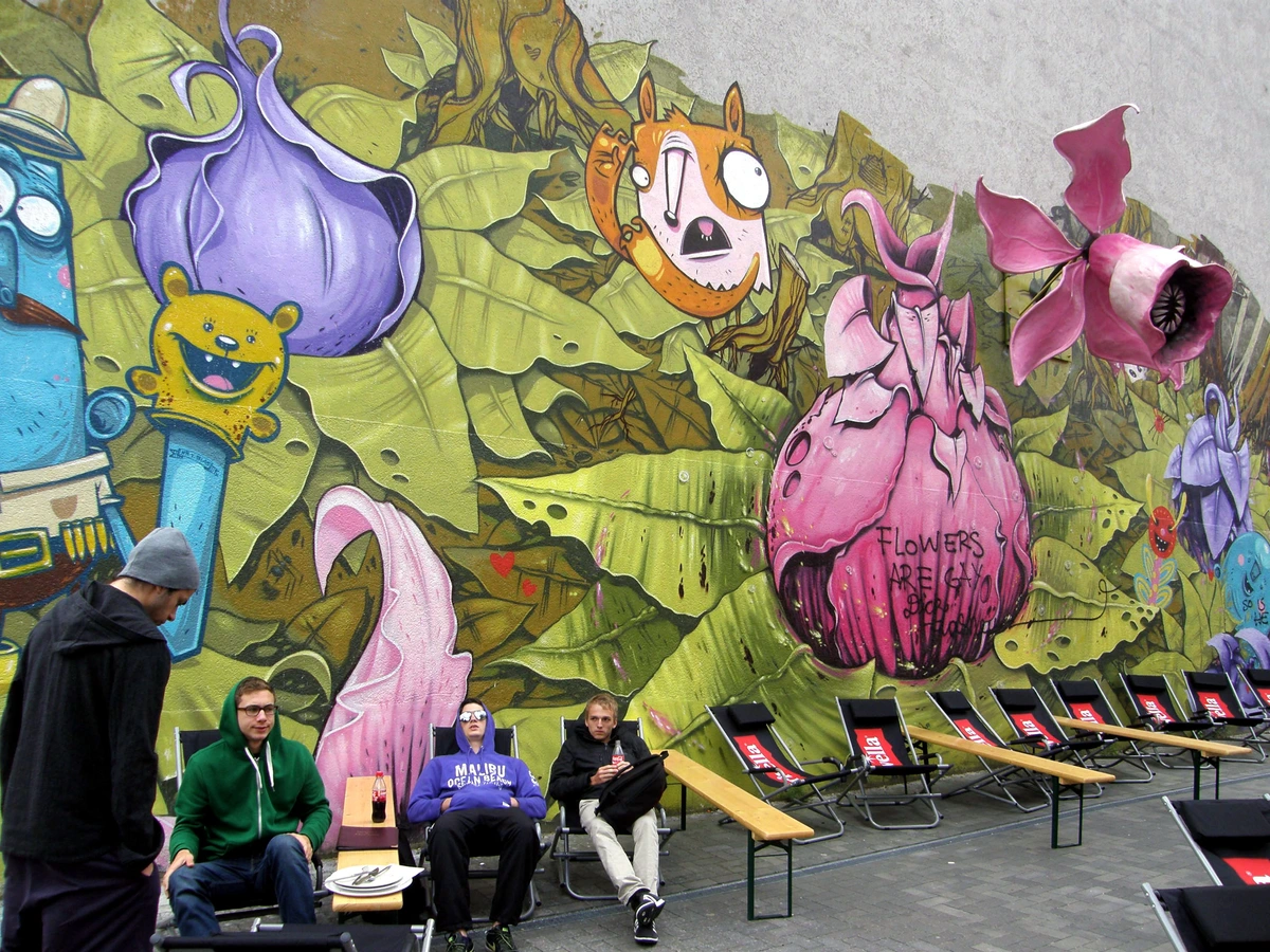 Vibrant graffiti mural in Berlin Kreuzberg featuring cartoon characters and flowers, with people sitting on lounge chairs in front.