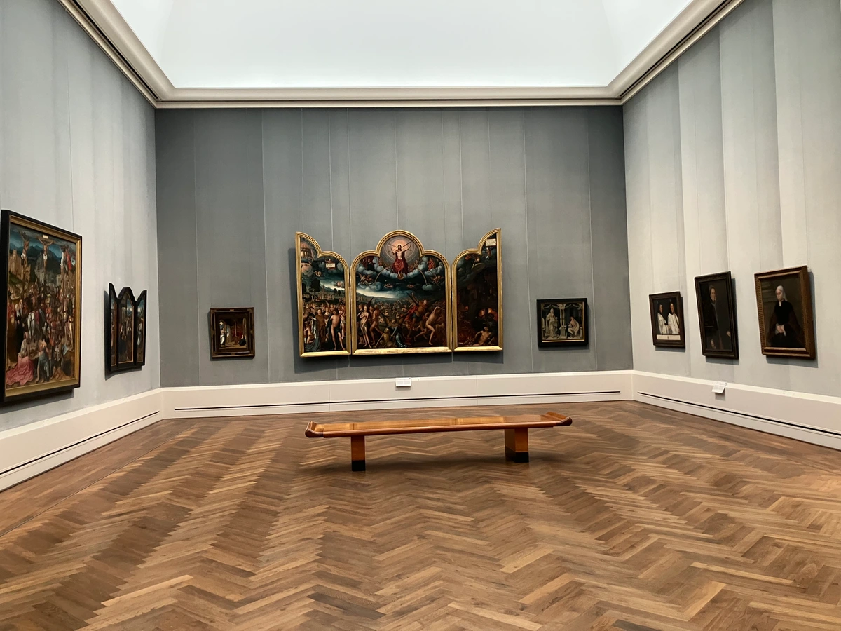 Interior view of Room VI at the Gemäldegalerie in Berlin, showcasing a collection of Old Master paintings, including a large triptych and several portraits, displayed on a grey wall with a wooden parquet floor and a central bench.