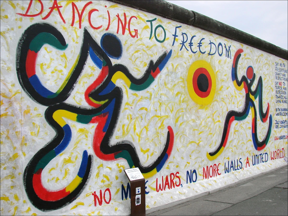 Mural on the East Side Gallery of the Berlin Wall depicting colorful figures dancing and the text 'Dancing to Freedom' and 'No More Wars, No More Walls, A United World'.