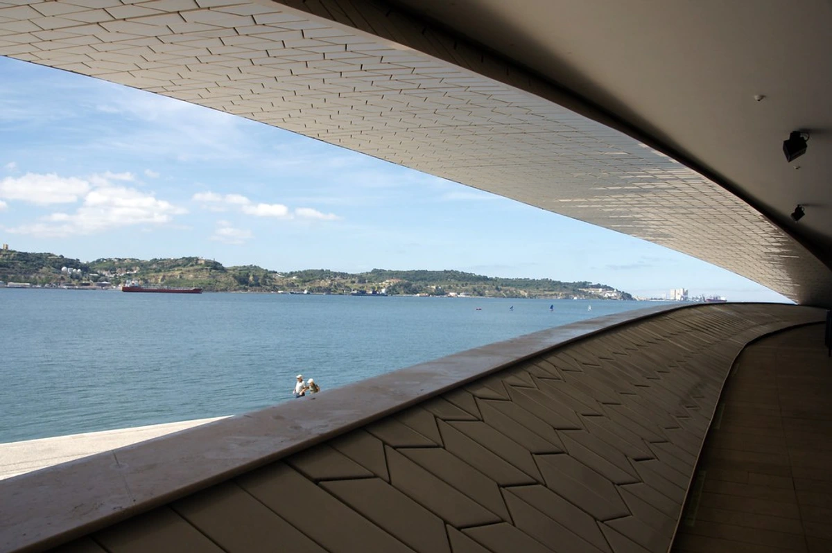 View of the Tagus River from the MAAT museum in Lisbon, Portugal, with a large cargo ship and sailboats on the water.