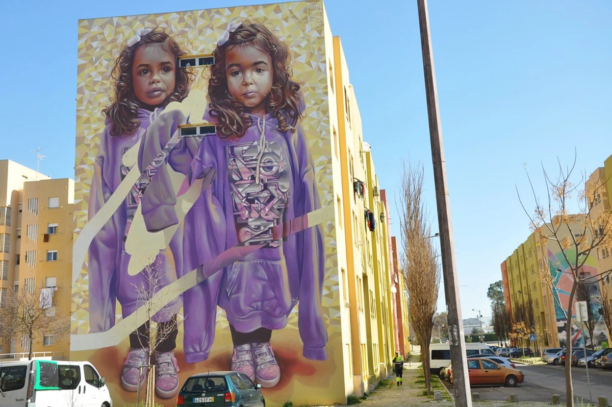 Vibrant street art mural of two young girls in purple hoodies, painted on the facade of a residential building in Bairro Padre Cruz, Lisbon, by artist Pariz One.
