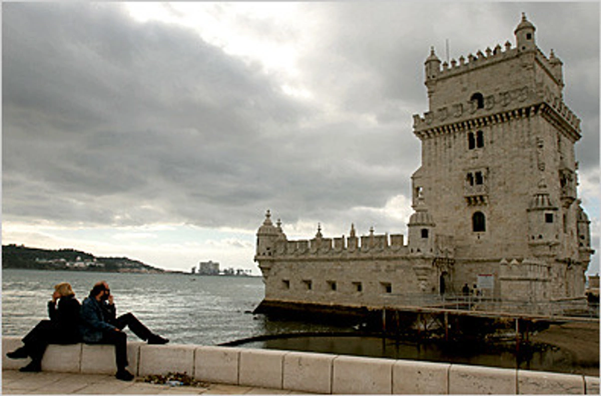 Belem Tower in Lisbon, Portugal, with two people sitting on a wall overlooking the Tagus River on a cloudy day.
