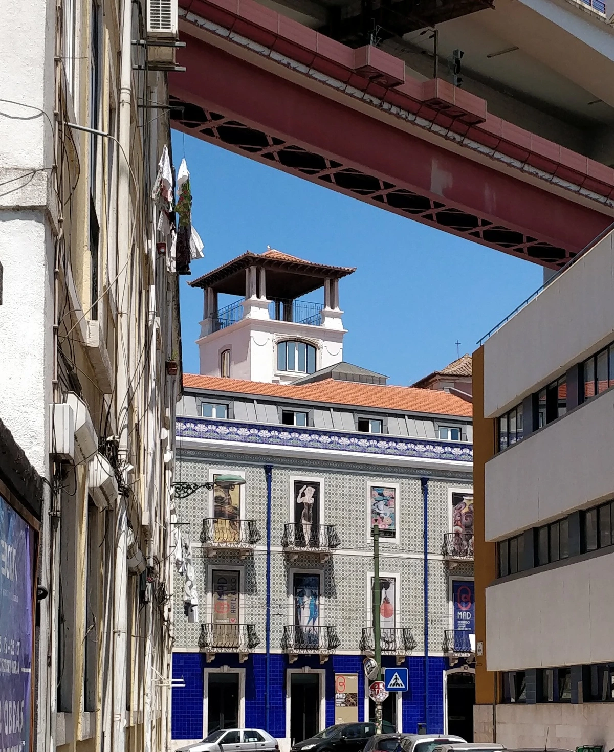 Facade of the B-MAD Museum in Lisbon, Portugal, featuring colorful azulejo tiles and balconies, with the iconic 25 de Abril Bridge visible overhead.