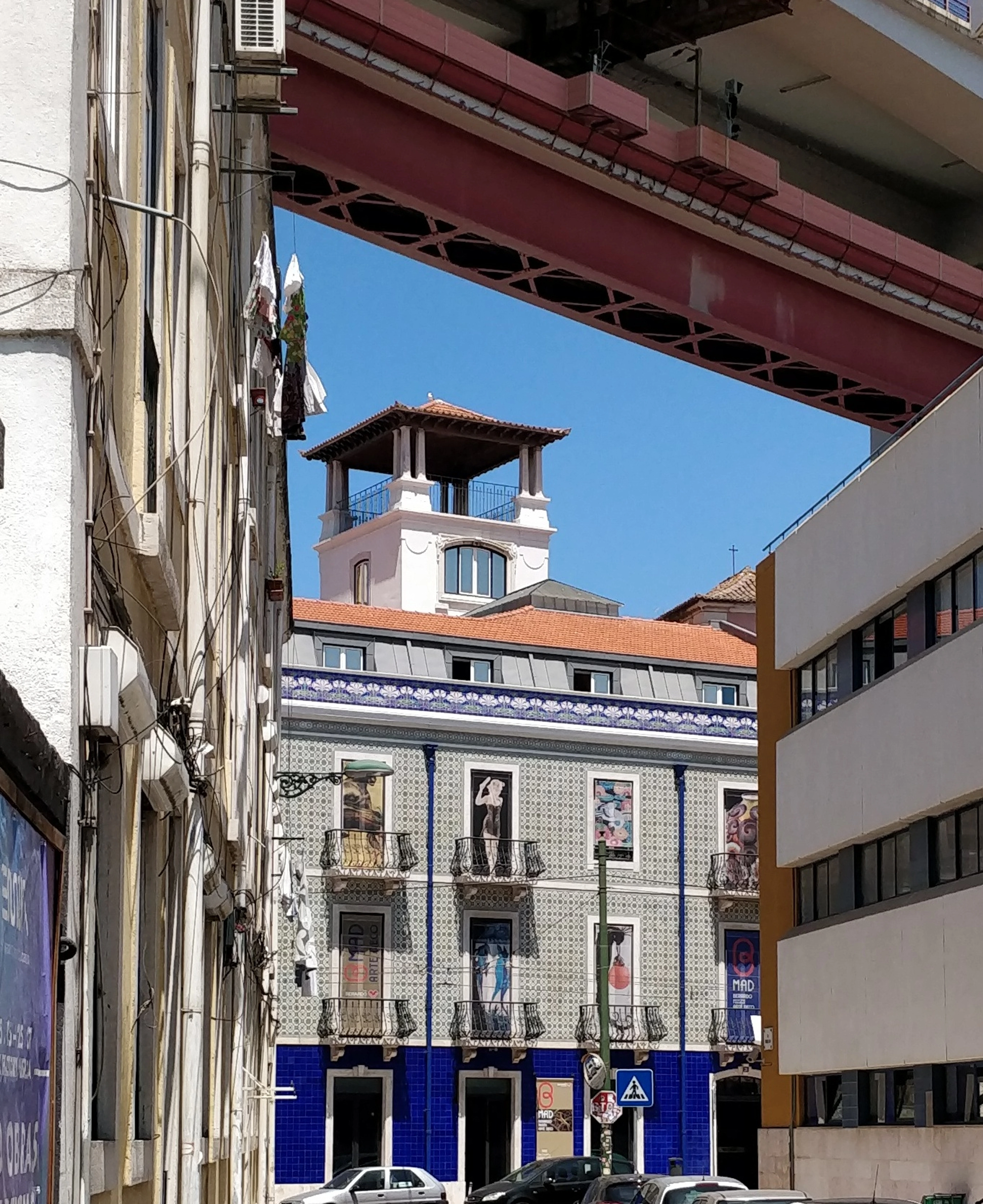 Facade of the B-MAD Museum in Lisbon, Portugal, featuring colorful azulejo tiles and balconies, with the iconic 25 de Abril Bridge visible overhead.