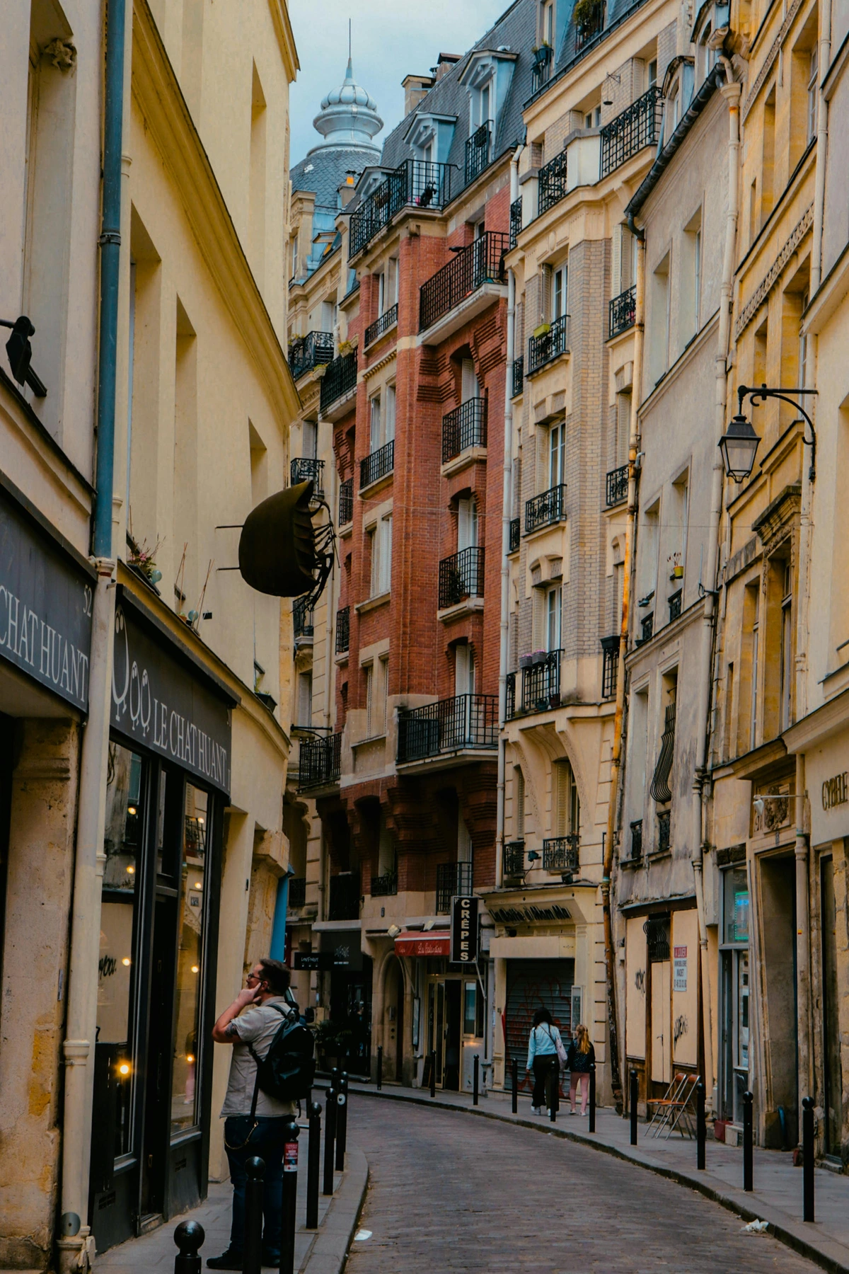 People walking on a cobblestone street between tall, narrow buildings with shops and balconies in Paris.