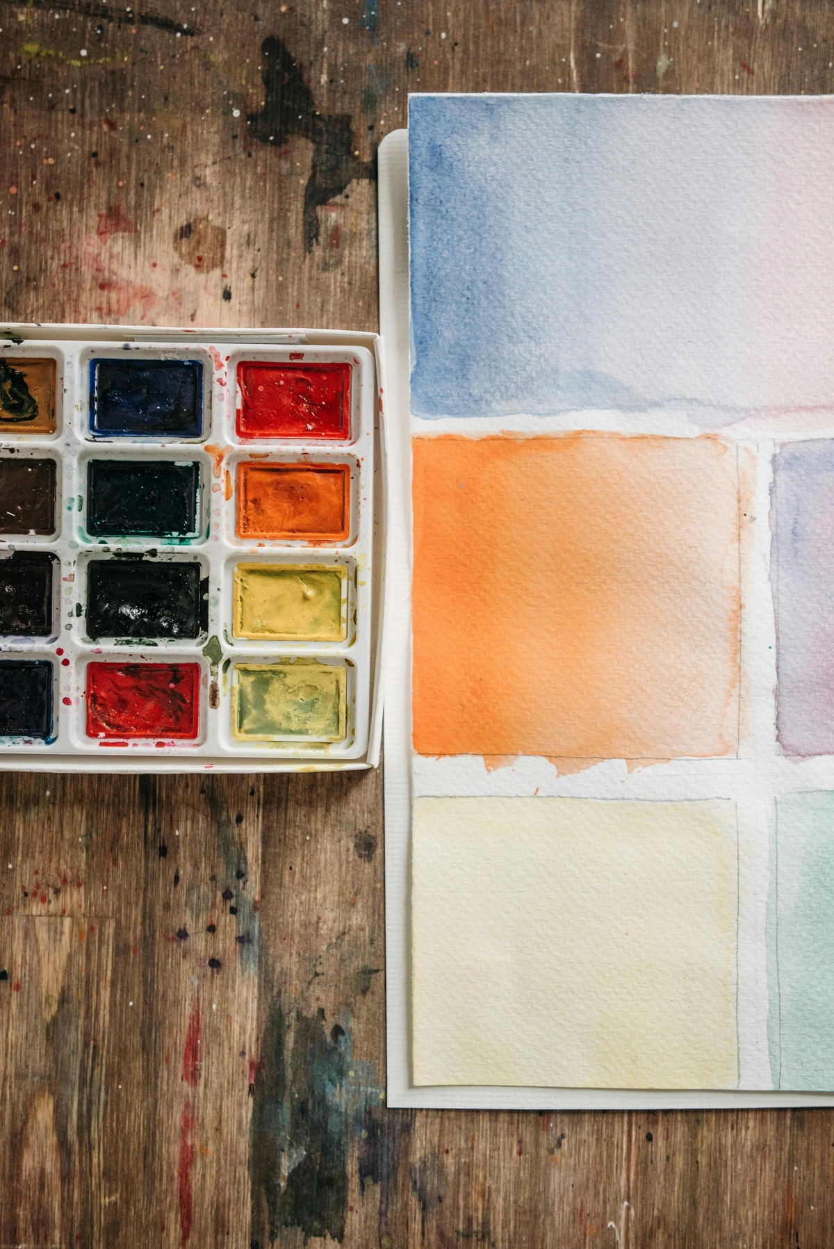 Close-up overhead view of a Winsor & Newton professional watercolor paint set with various colors on a rustic wooden surface with paint splatters.