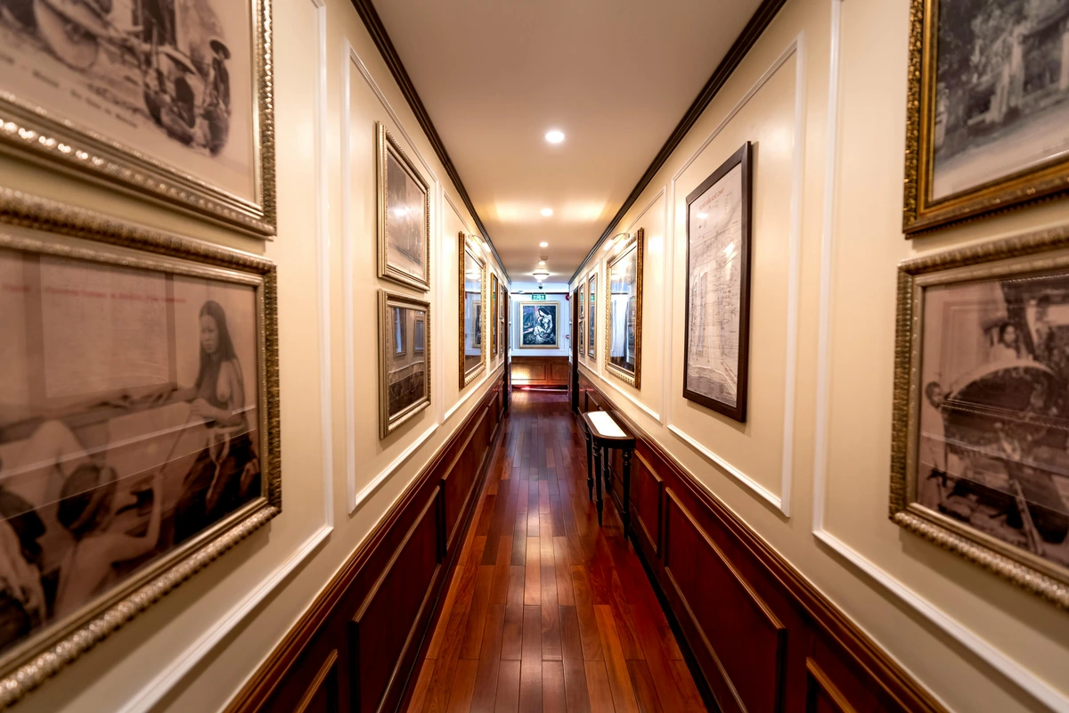 Long, narrow cruise ship hallway decorated with framed art and paintings on the walls, featuring polished wood paneling and a gleaming hardwood floor.