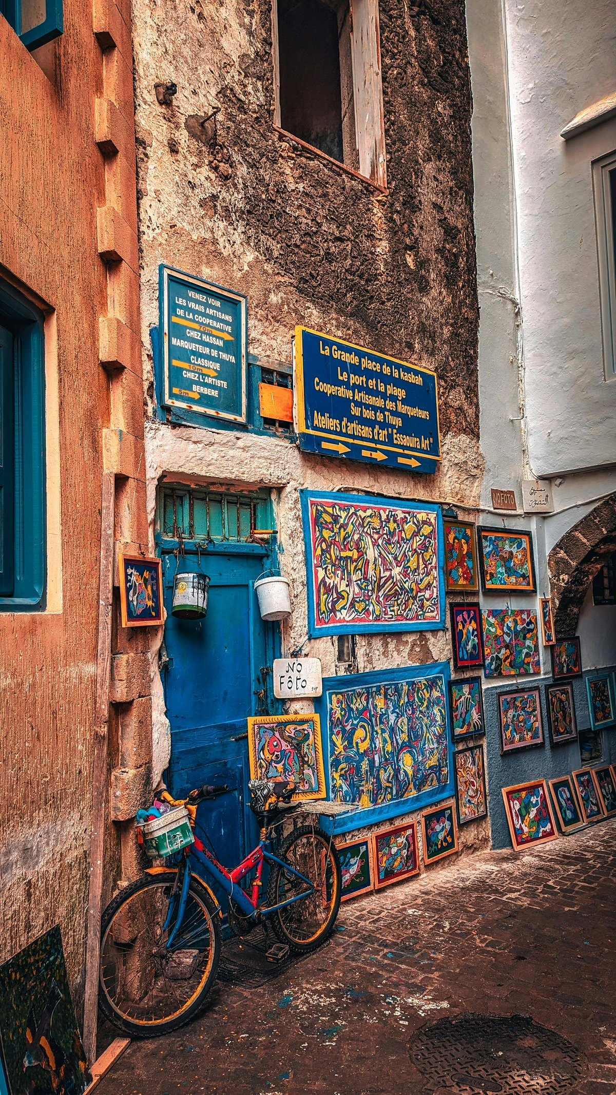 Colorful art gallery alleyway in Essaouira, Morocco, featuring vibrant paintings displayed along a narrow, textured hallway with a blue door and a bicycle.