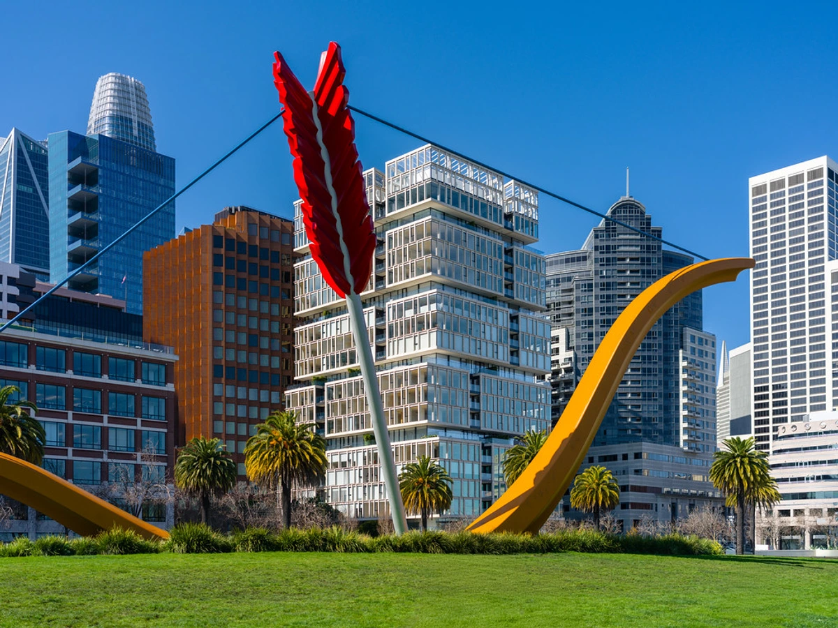 Cupid's Span Sculpture Art Installation on Embarcadero San Francisco, a striking red arch installation attracting a wide audience, in a lush green park with palm trees, city skyscrapers, and clear blue skies visible in the background