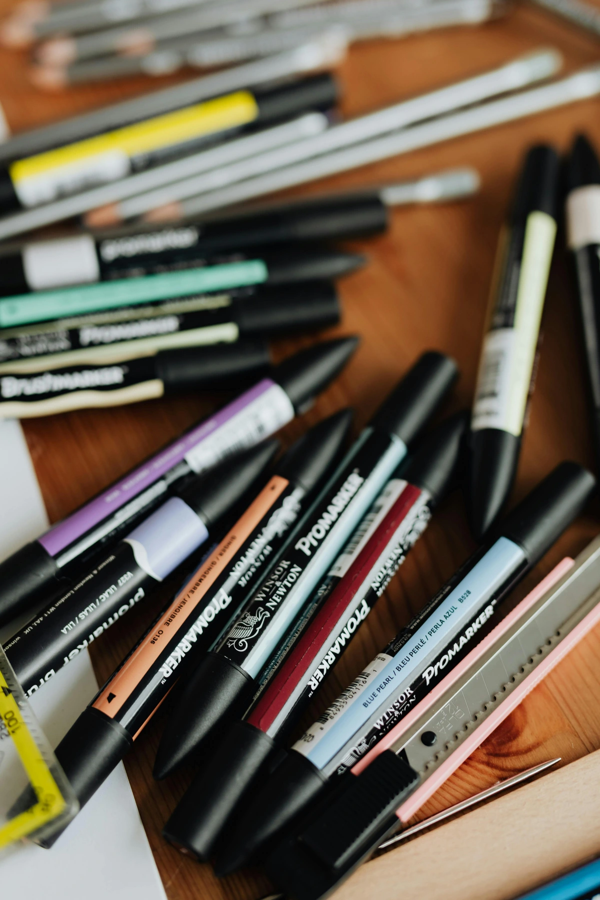 A collection of Winsor & Newton Promarker alcohol-based markers in various colors, along with pencils and a craft knife, arranged on a wooden surface, ideal for artists. | Een verzameling Winsor & Newton Promarker alcoholmarkers in verschillende kleuren, samen met potloden en een hobbymes, gerangschikt op een houten ondergrond, ideaal voor kunstenaars.