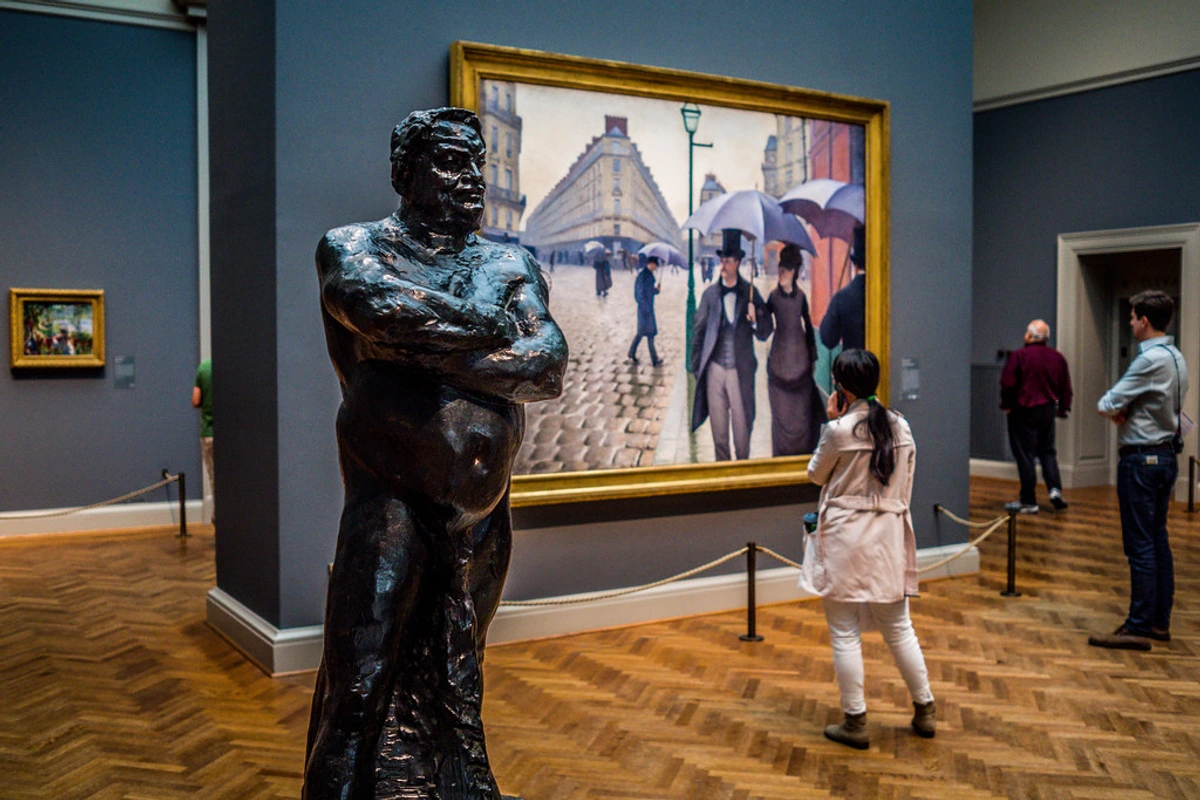 A bronze statue of a man with his arms crossed stands in the foreground at the Art Institute of Chicago, with a painting of a Parisian street scene and museum visitors in the background.