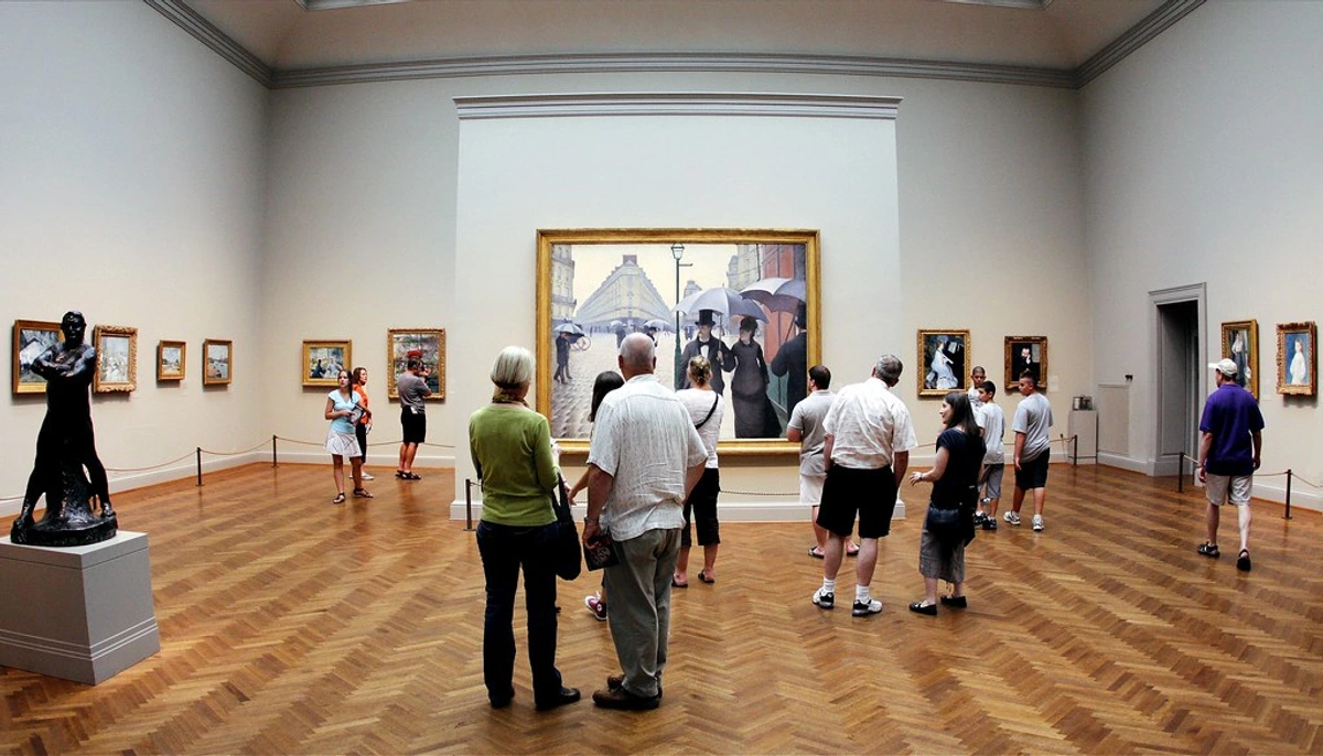 Visitors admiring paintings and a sculpture in a well-lit art museum gallery with a parquet floor.