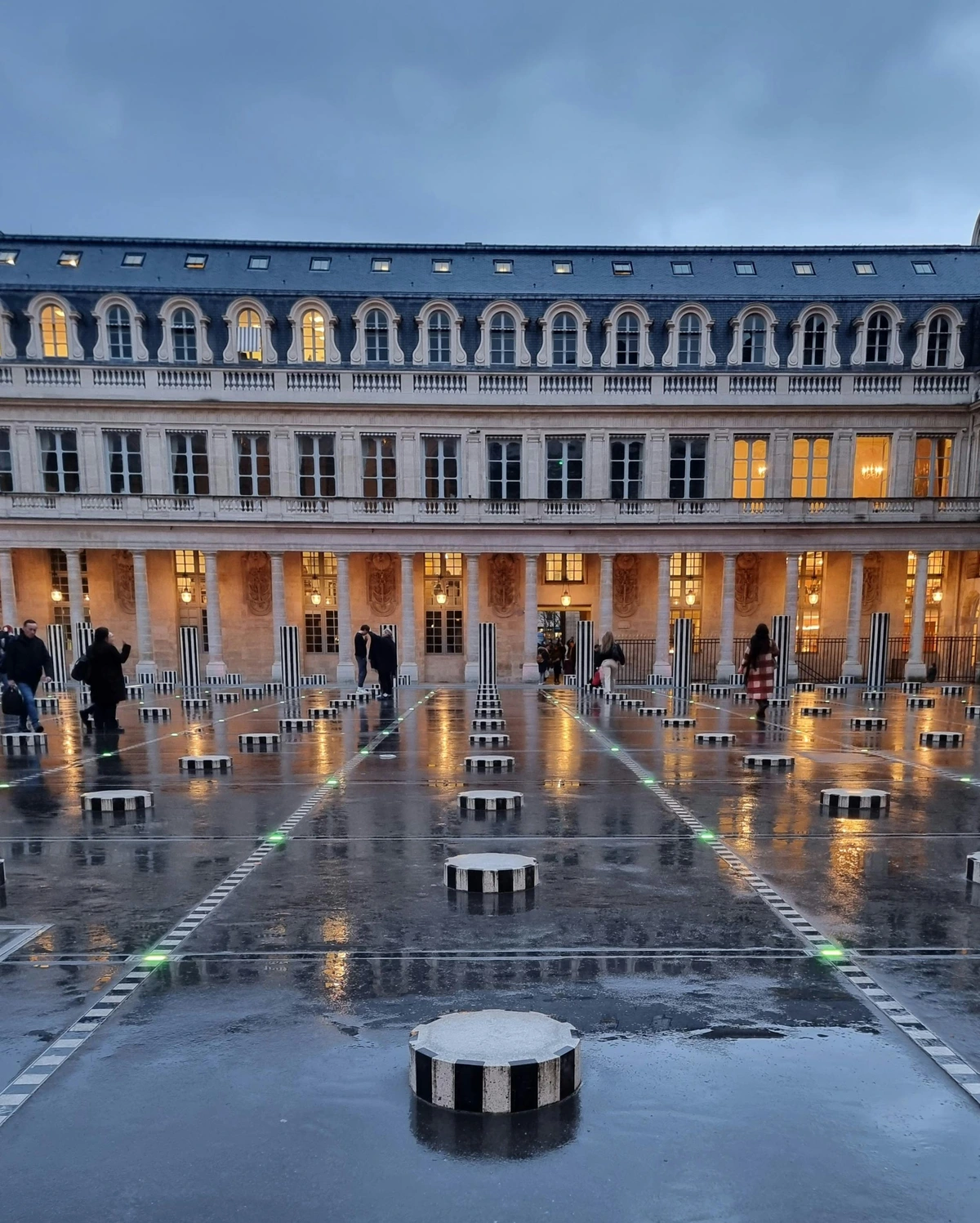 Tourists exploring the Louvre Museum entrance with iconic glass pyramid and black-and-white striped installations under evening illumination.