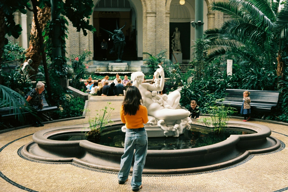 Woman admiring modern art installation in indoor courtyard with blooming Rhodian marble sculpture and academic discussion in background museum pavilion