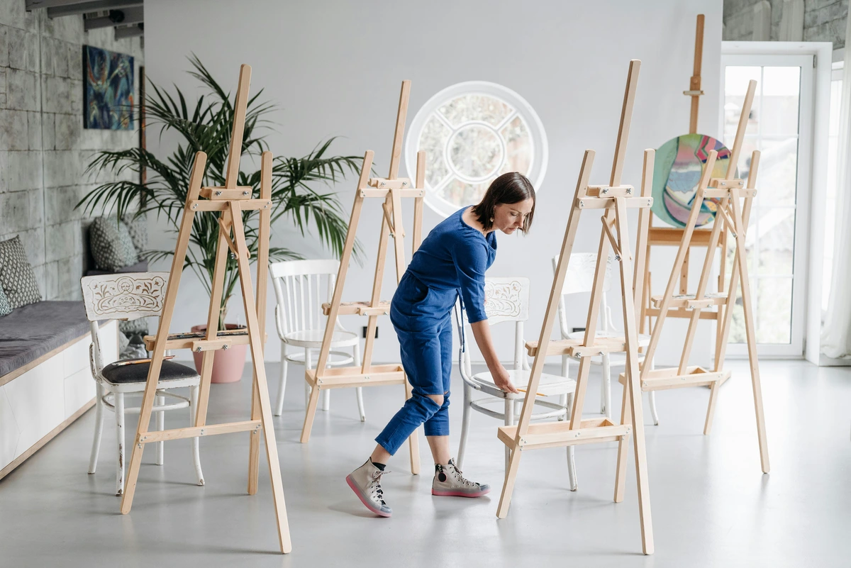 A woman in a blue jumpsuit inspects wooden easels in a bright, spacious art studio with large windows and plants.