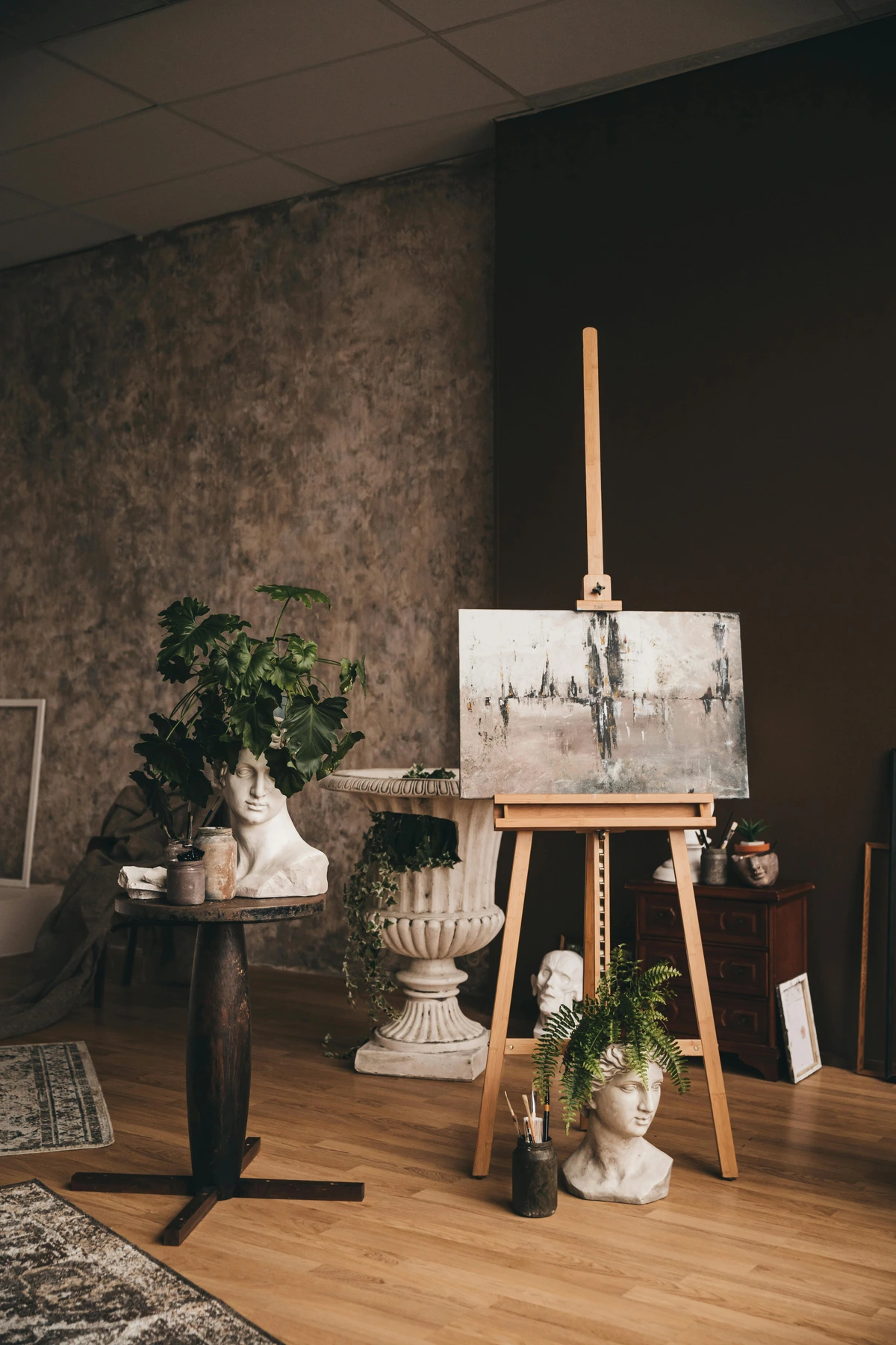 Painting on a wooden easel with classical bust sculptures and plants in an art studio setting.