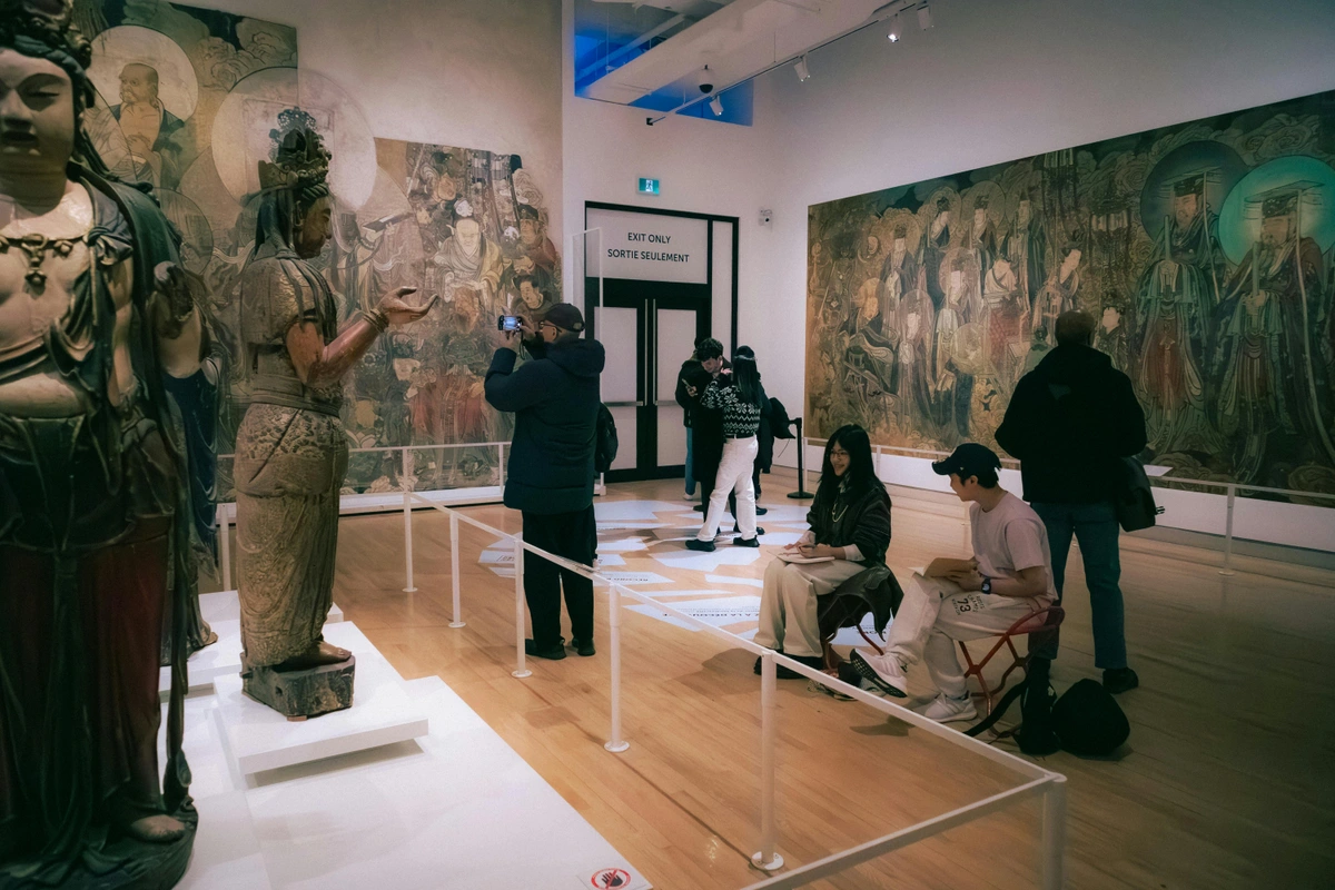 Visitors admire ancient Asian art, including large statues and detailed murals, within a brightly lit museum in Toronto, Canada.