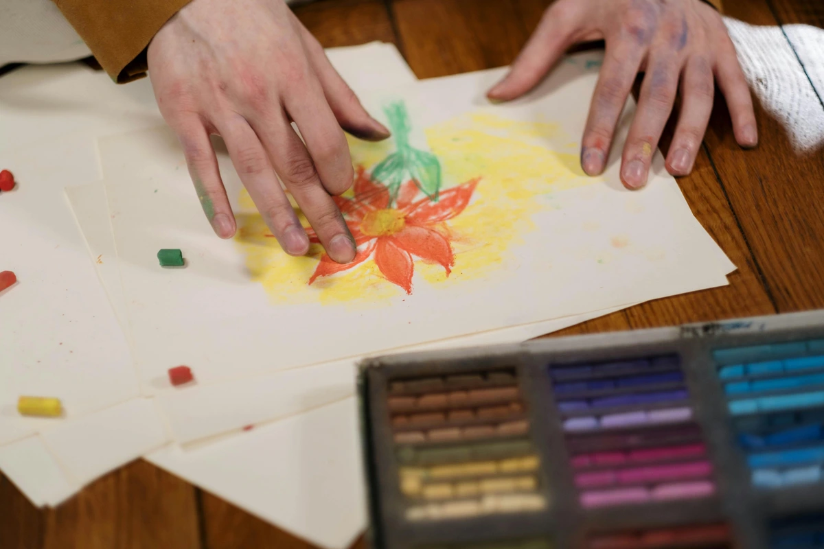 Close-up of a person's hands drawing a flower with colorful chalk pastels on paper.