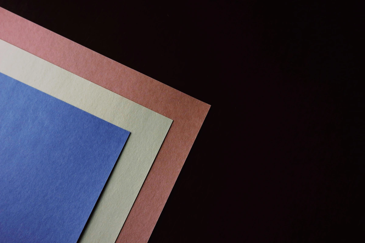 Close-up of layered pastel drawing papers in blue, beige, and pink on a black surface, showcasing texture.