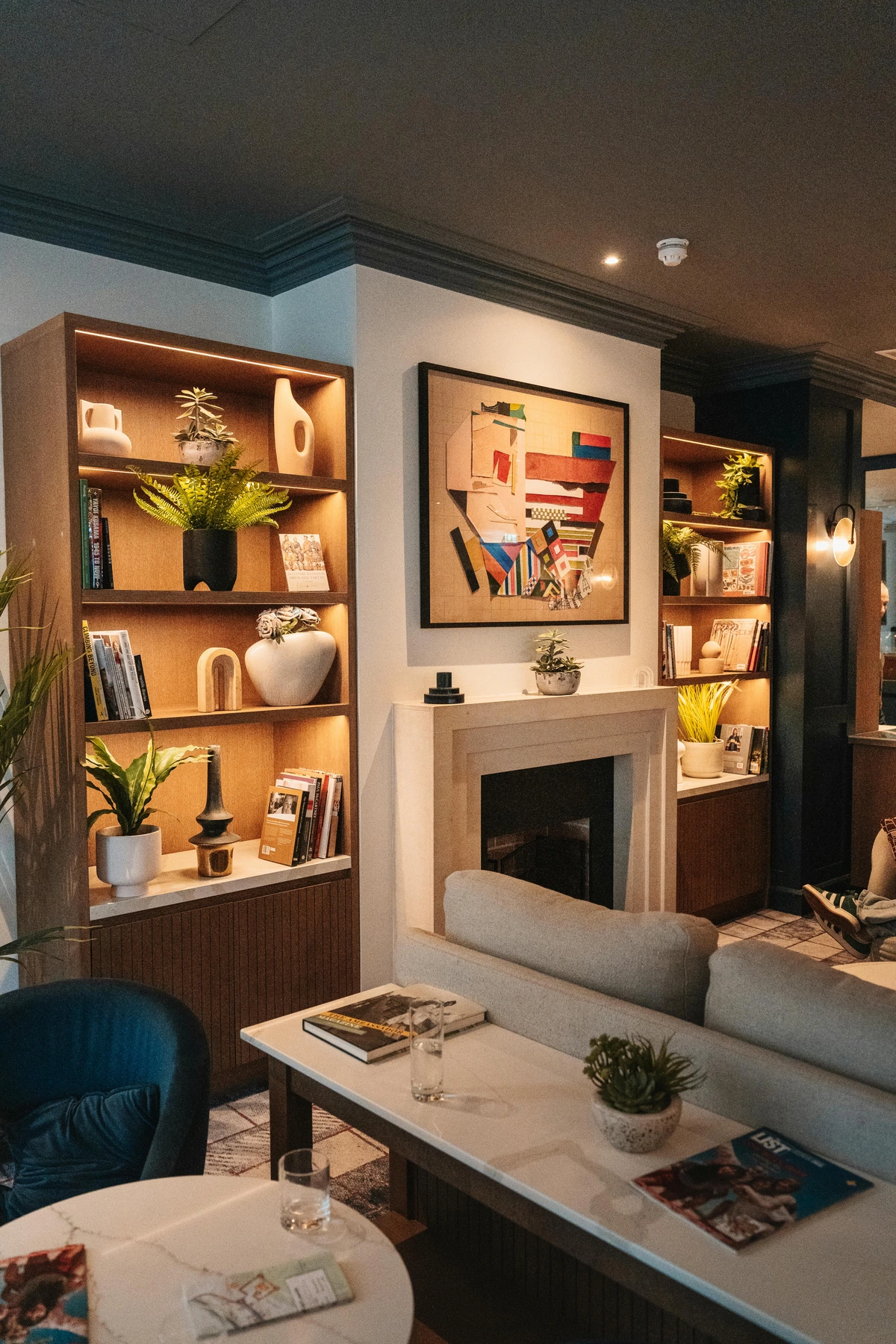 Modern cozy living room featuring a fireplace with abstract art above, flanked by illuminated bookshelves and comfortable seating.