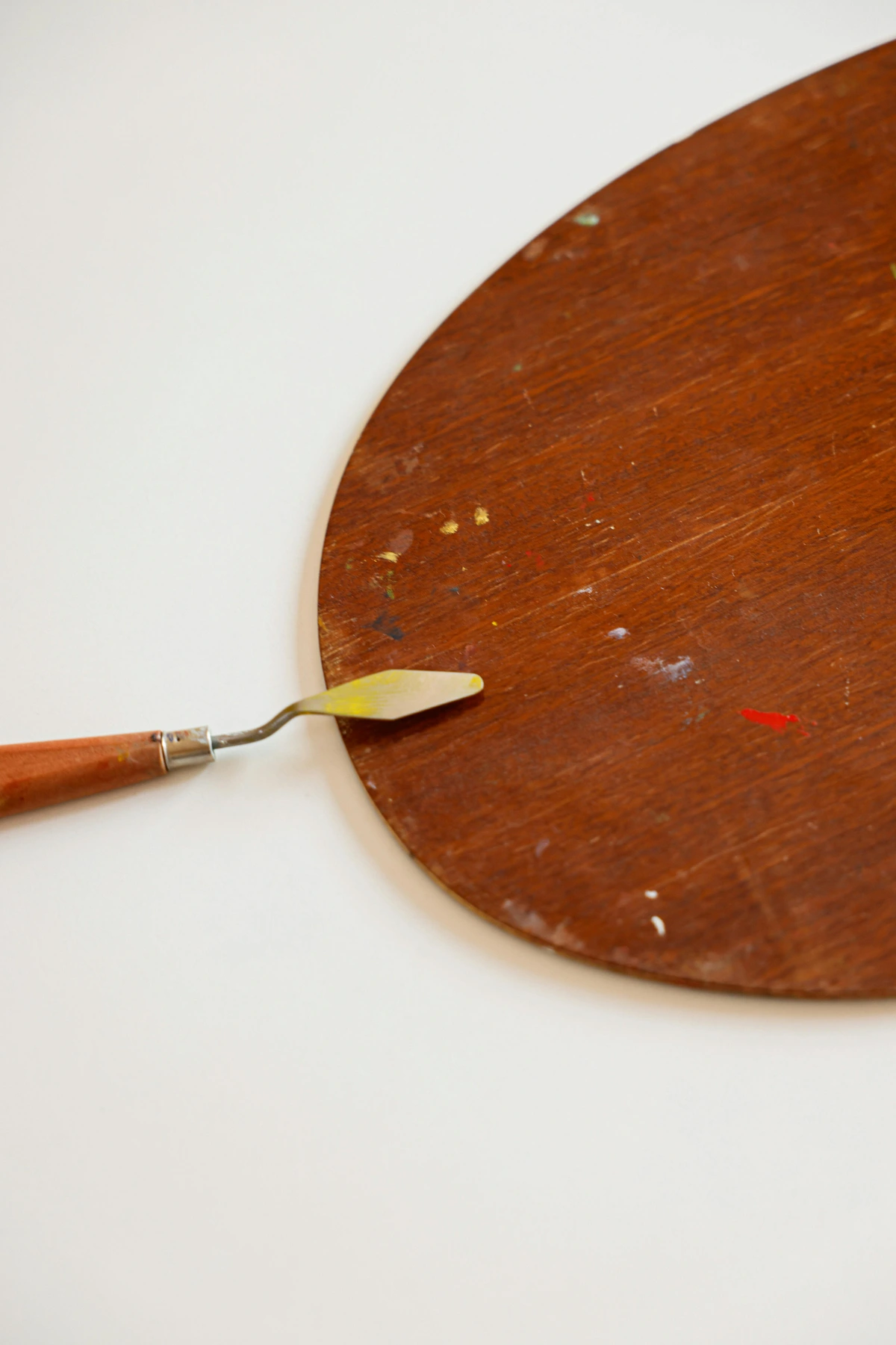 A palette knife with a yellow tip rests on a wooden artist's color mixing palette, which has small specks of paint on its surface.