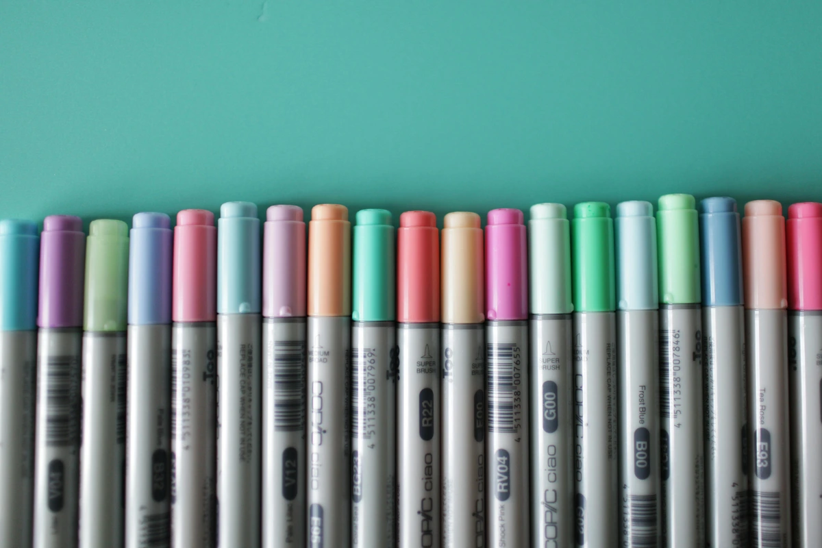Assorted color Copic markers arranged in a row on a teal background, ideal for professional artist reviews.