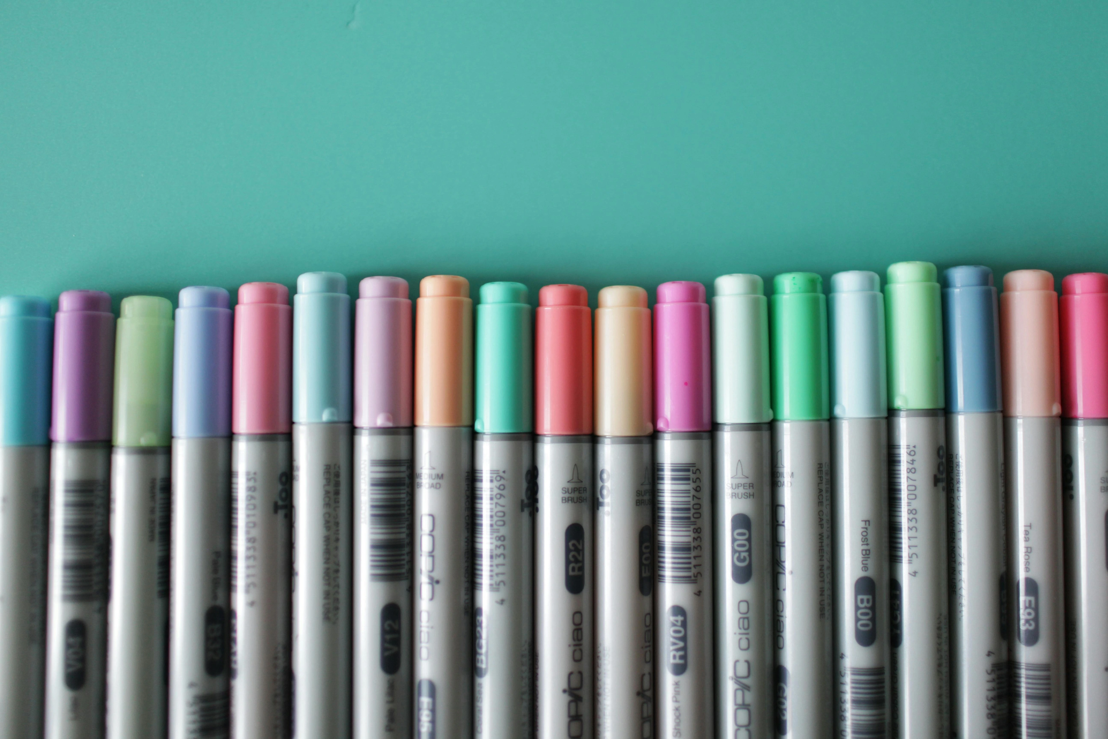 Assorted color Copic markers arranged in a row on a teal background, ideal for professional artist reviews.