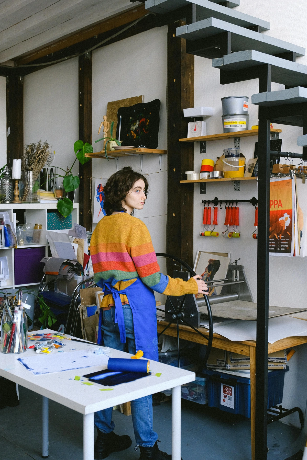 Artist operating printing machine in modern workshop with art supplies and tools visible.