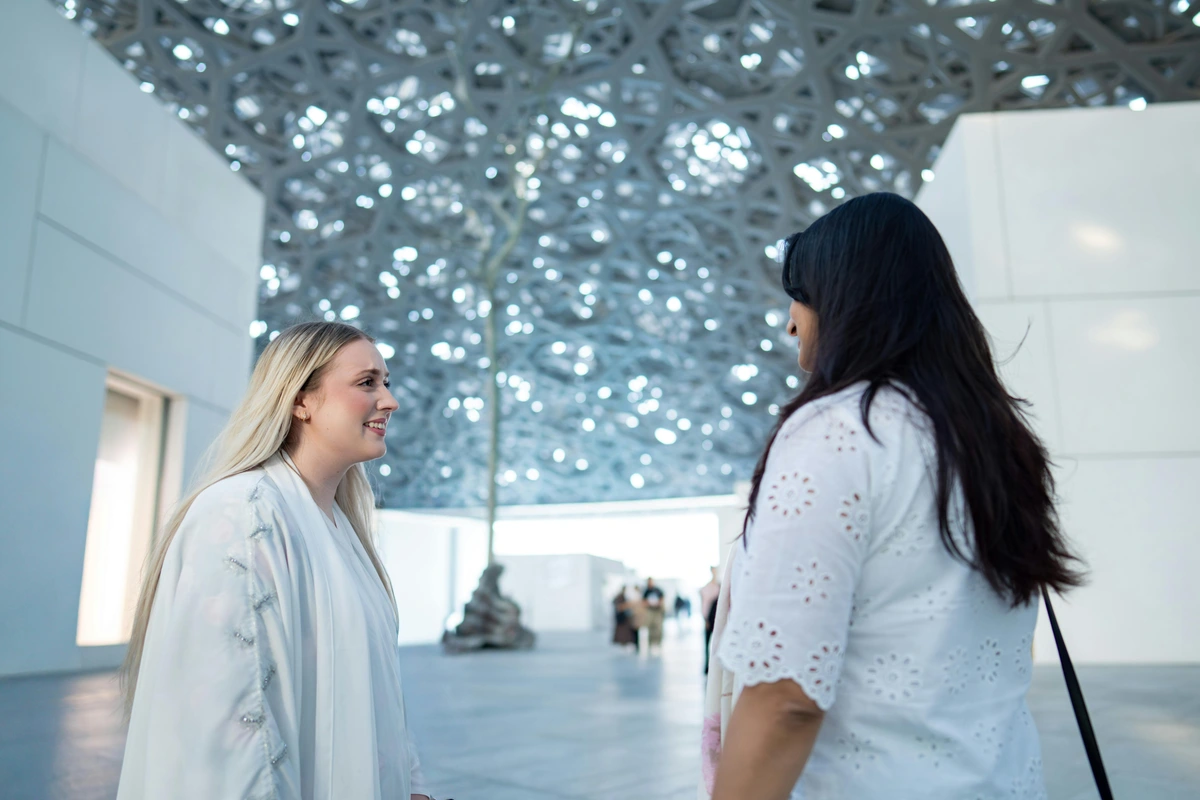 Two women engaged in conversation under the iconic Louvre Abu Dhabi dome, symbolizing intellectual curiosity.