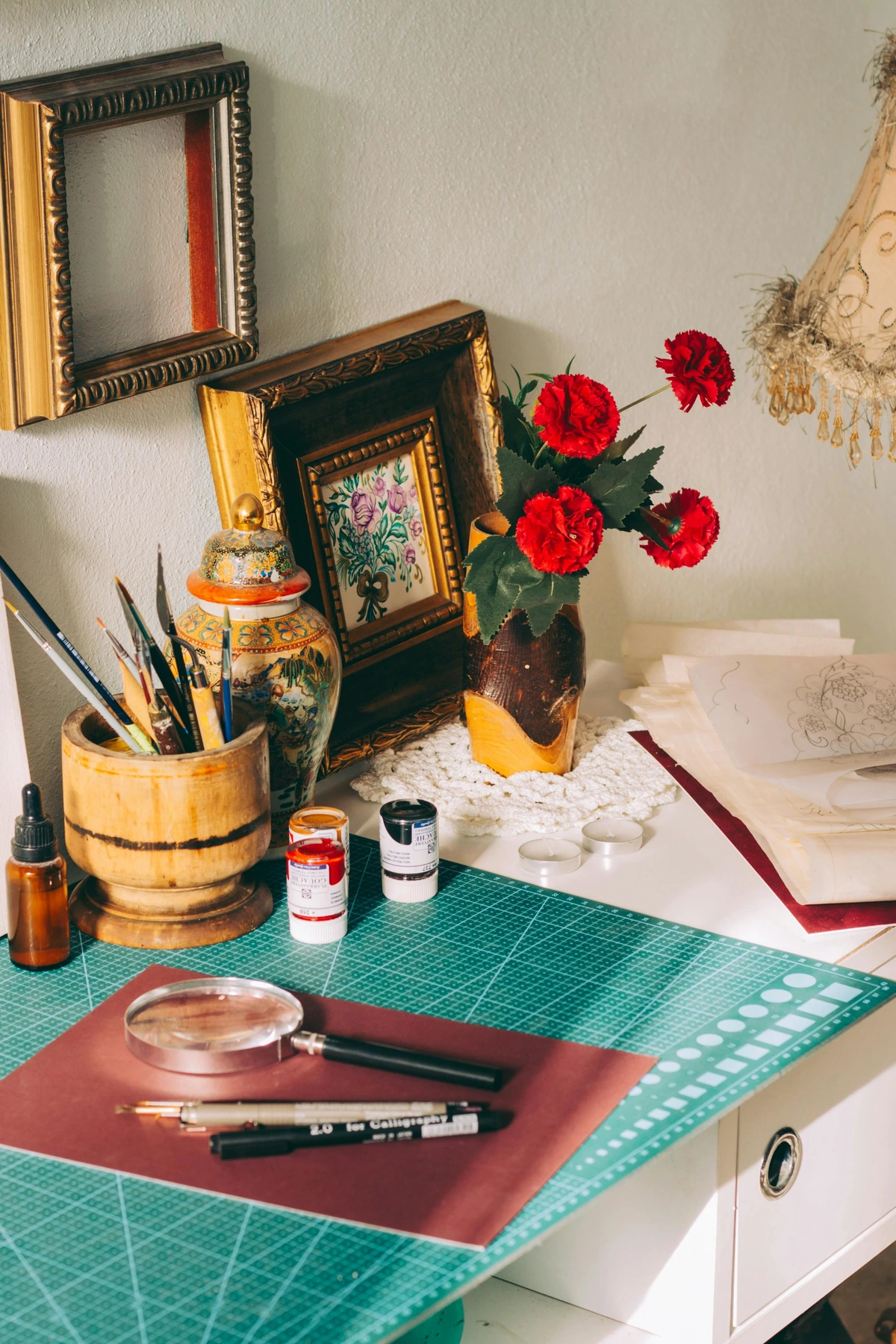 Vintage art studio workspace surrounded by red carnations and art supplies on a green cutting mat with painting tools, magnifying glass, and colorful paint tubes nearby, capturing the creative process and artistic craftsmanship in a classic french country setting with wooden countertop and gray walls in the background