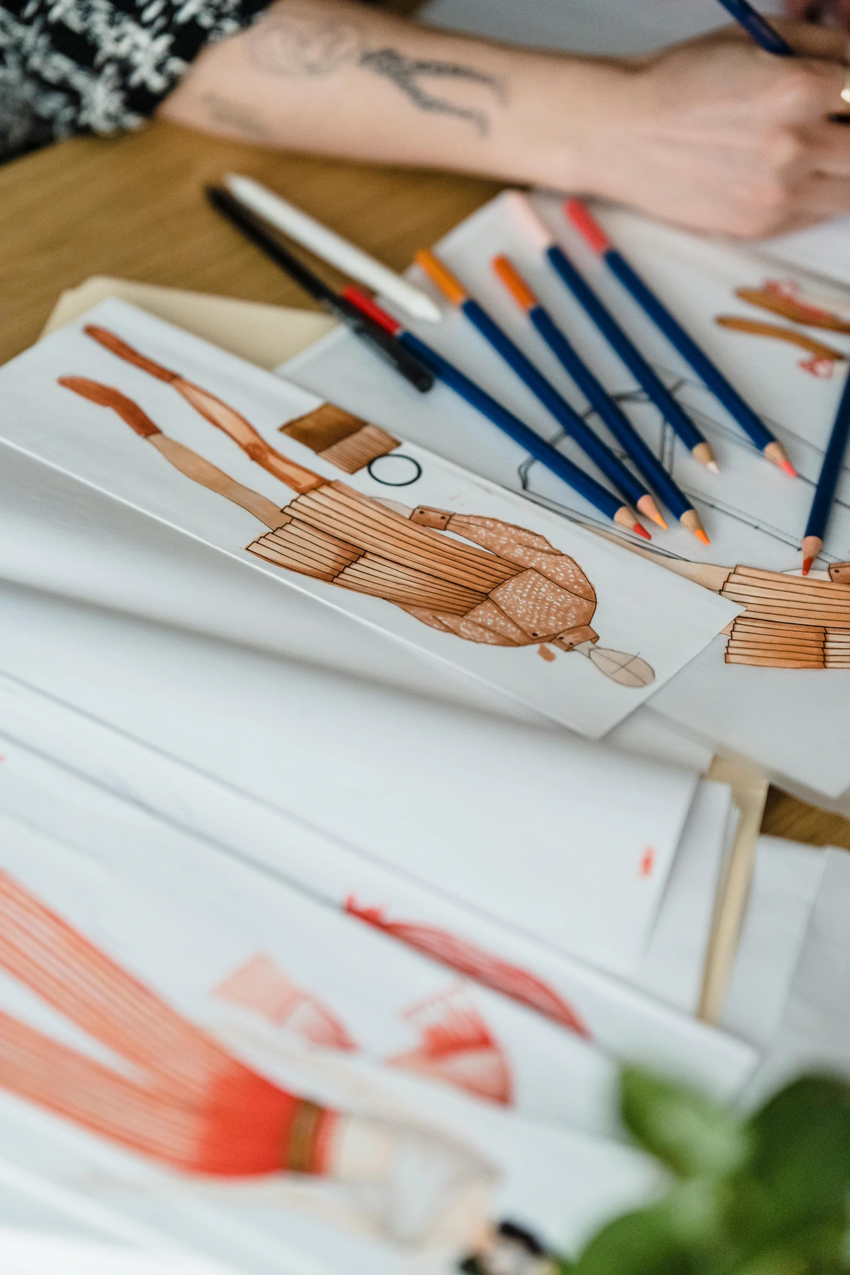 Illustrator sketching clothing designs on paper at a table with artistic tools and detailed fabric textures visible in creative workspace setting
