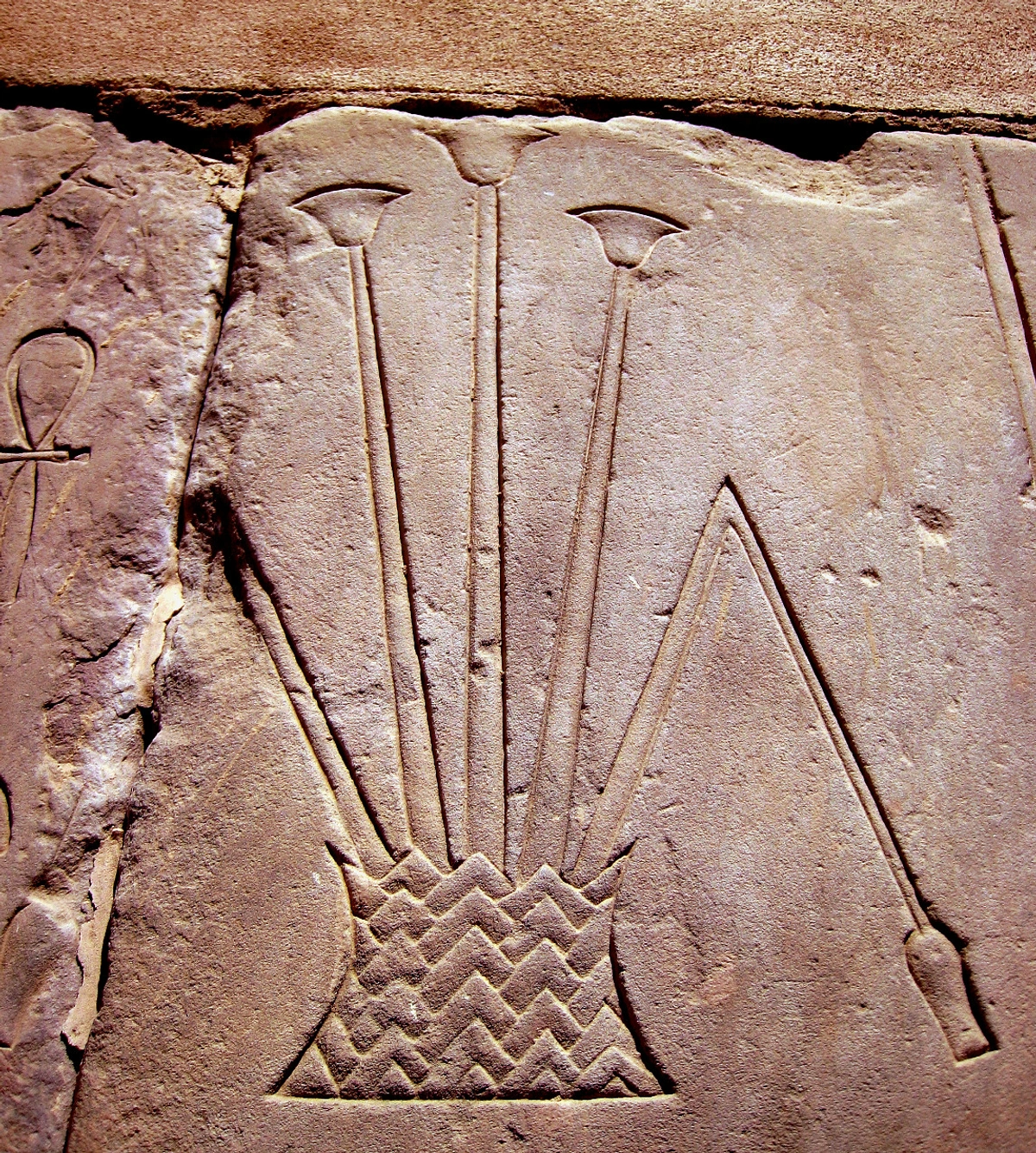 Ancient Egyptian relief carving of papyrus flowers in a basket, with an ankh symbol to the left.