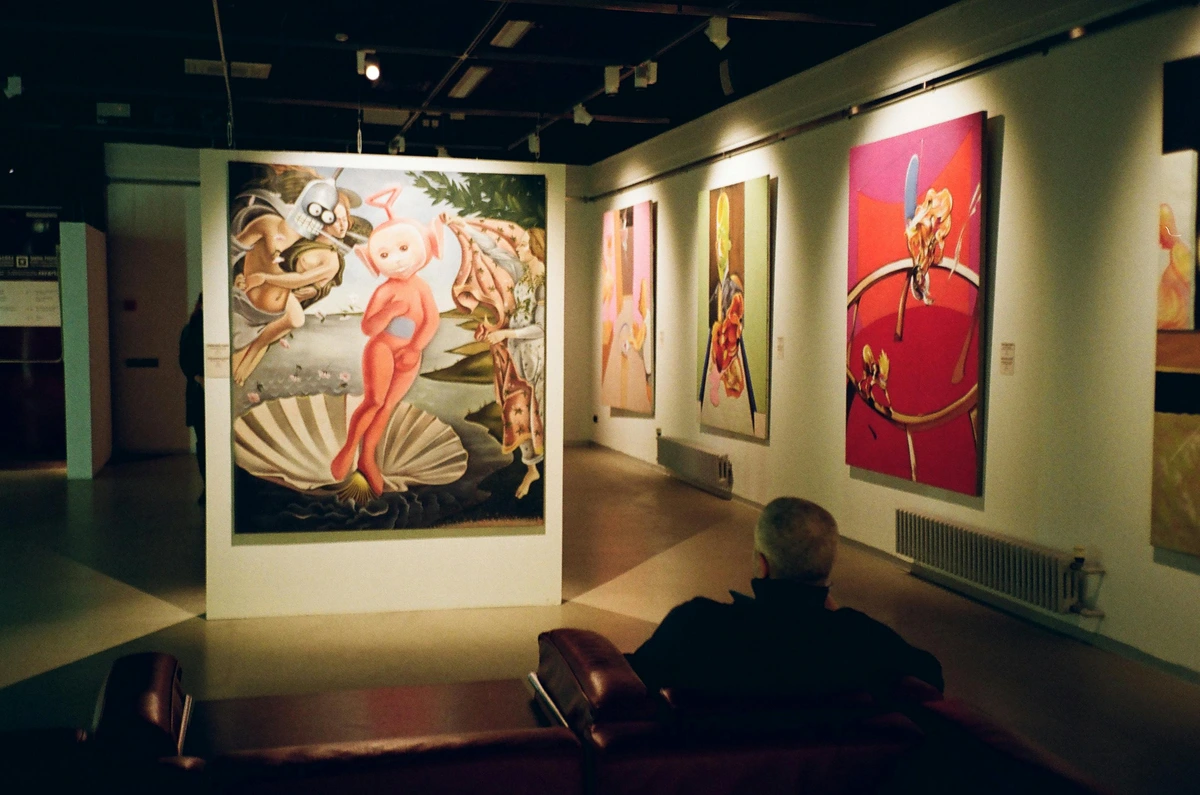 A man seated in a room surrounded by diverse paintings at a free art museum, observing modern and classical artworks from a side perspective.