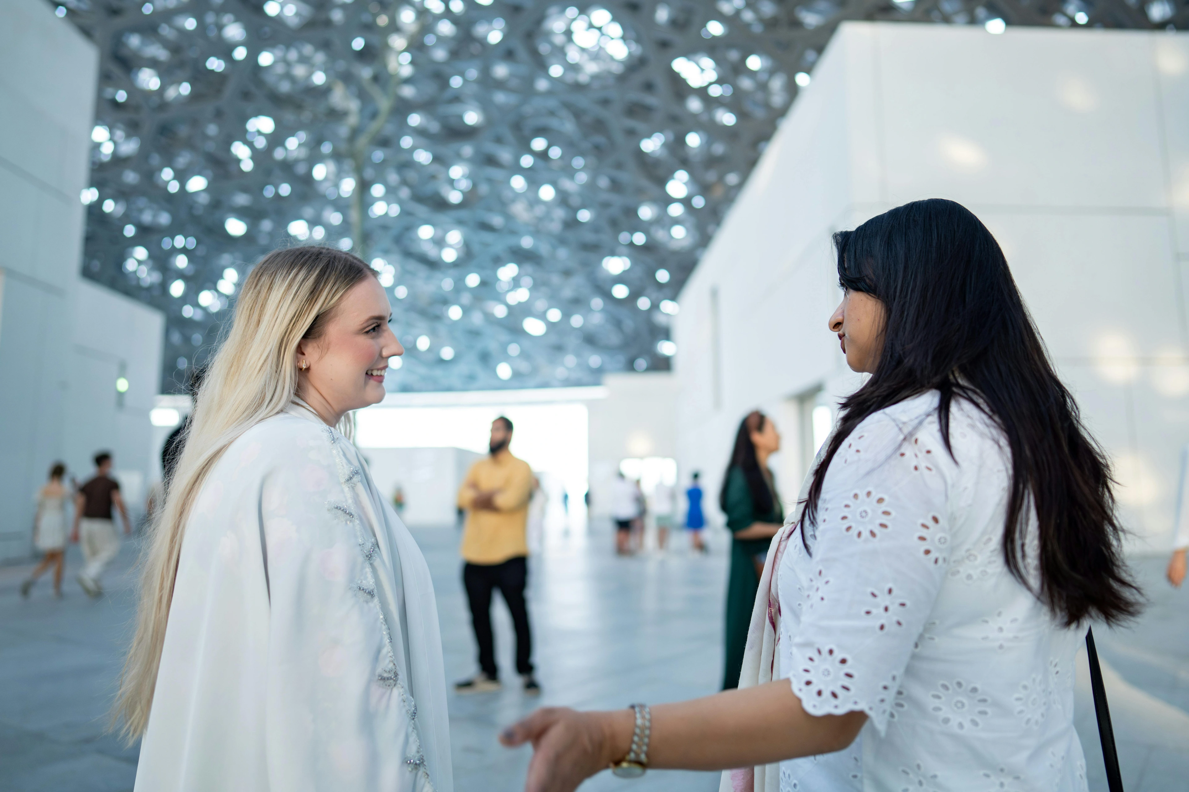 Two women engaged in conversation within a modern art museum interior featuring an intricate metal ceiling structure.