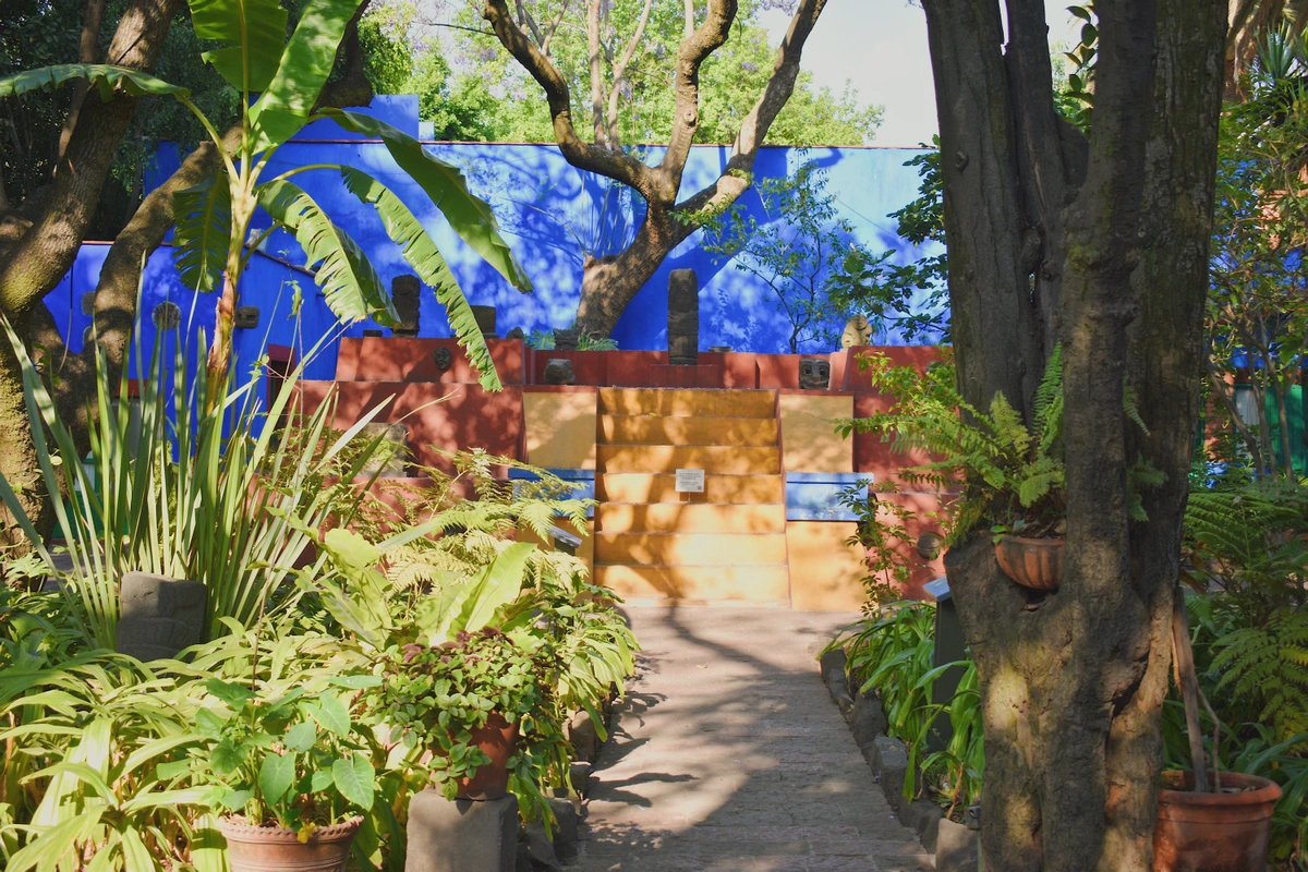 Lush garden path leading towards the vibrant blue walls of the Frida Kahlo Museum in Coyoacan, Mexico City, with tropical plants and ancient artifacts.