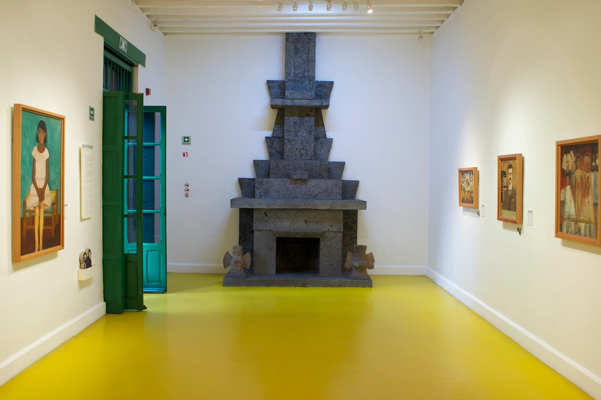 Interior of Room 1 at the Frida Kahlo Museum (Casa Azul) in Coyoacan, Mexico City, featuring a large stone fireplace, paintings on the wall, and a bright yellow floor.