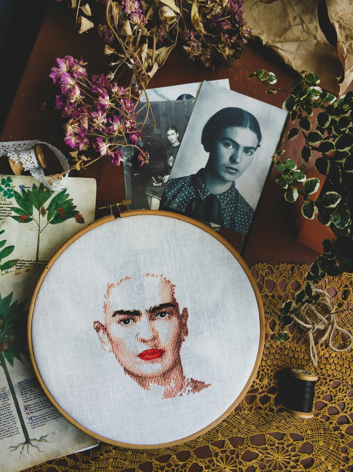 Frida Kahlo cross-stitch portrait in an embroidery hoop, surrounded by vintage photos, dried flowers, and botanical illustrations.